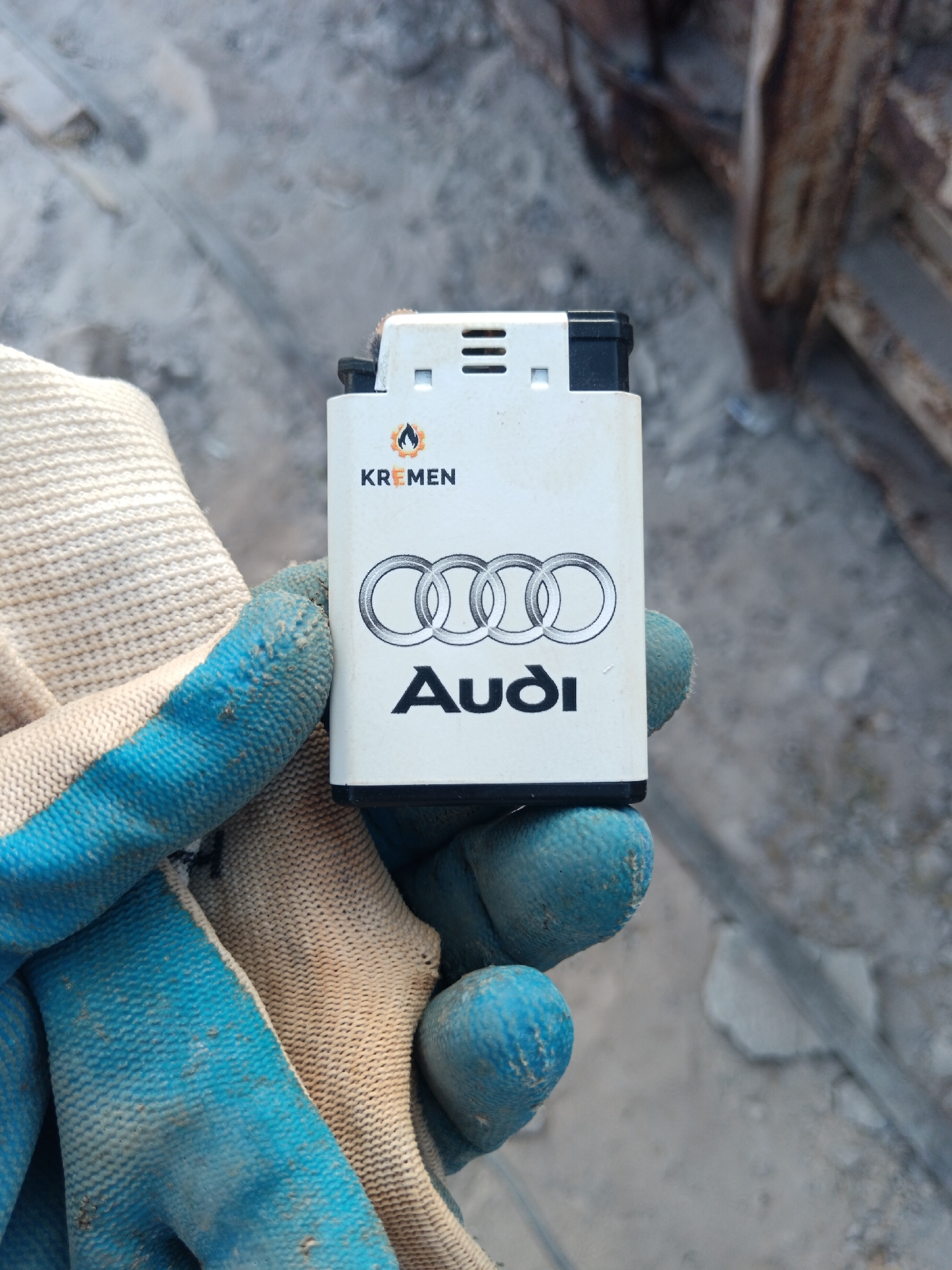 In Aragatsavan, Armenia, a close-up view shows a hand wearing a worn, light-colored knitted glove with blue rubberized fingertips, holding a white disposable lighter. The lighter features the Audi rings logo in black and the word "Audi" beneath it. Above the Audi logo is a smaller logo with a flame and the word "KREMEN". The background is blurred and appears to be an outdoor, urban environment with concrete surfaces and a rusted metal structure.