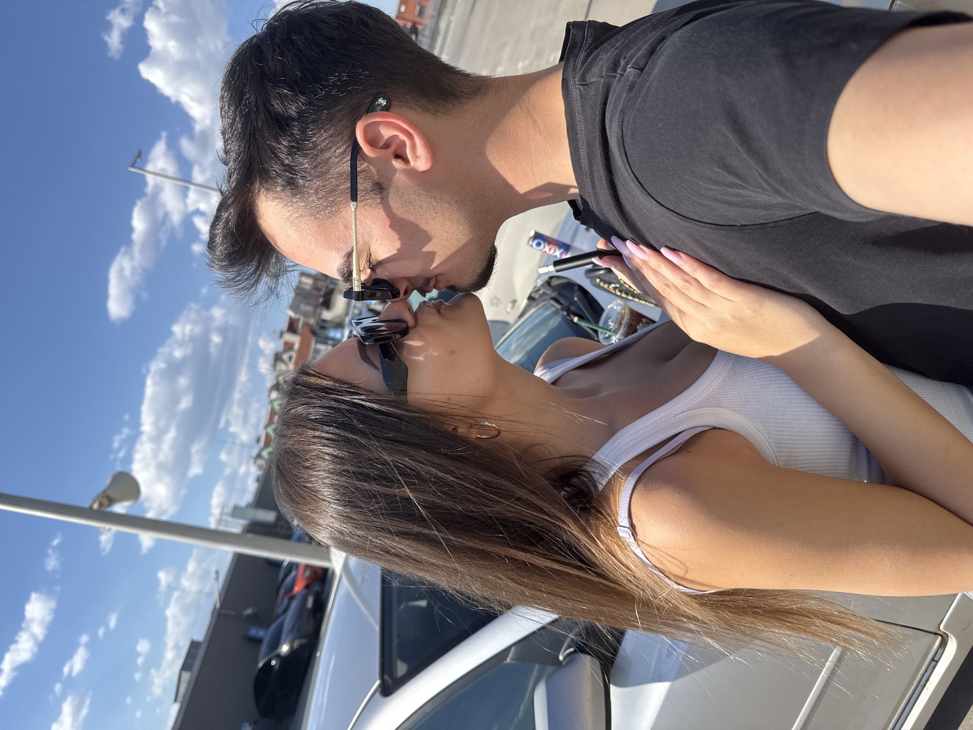 A man and a woman are kissing. The man has short dark hair, a black t-shirt, and gold-rimmed sunglasses. The woman has long brown hair and is wearing a white tank top and black sunglasses. They are positioned in front of a silver car. In the background, there's a sky with scattered clouds and what appears to be a building. There is an iced coffee drink sitting on the car along with a vape pen and a pack of "OXXI" brand cigarettes. I can't be certain about the location being Sofia, Bulgaria based on this single image. I can only describe what is visible.