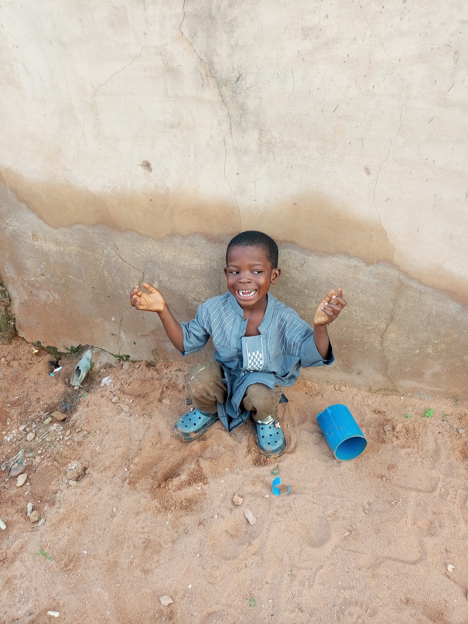 A young boy is crouching on sandy ground in front of a textured, light-colored wall with brown staining. He is wearing a gray, patterned tunic and brown checkered pants, with blue clogs on his feet. His hands are raised, palms facing outward, and he has a wide, toothy grin. To his right, there is a blue plastic cylindrical object on the ground. To his left, there is some debris including a broken piece of plastic and a small, greenish-gray object. The environment appears to be outdoors, with a rough, uneven surface of sand and scattered small rocks and bits of debris. The wall behind him shows signs of wear and possibly water damage. The lighting suggests it is daytime. The location is identified as Tempure, Nigeria.