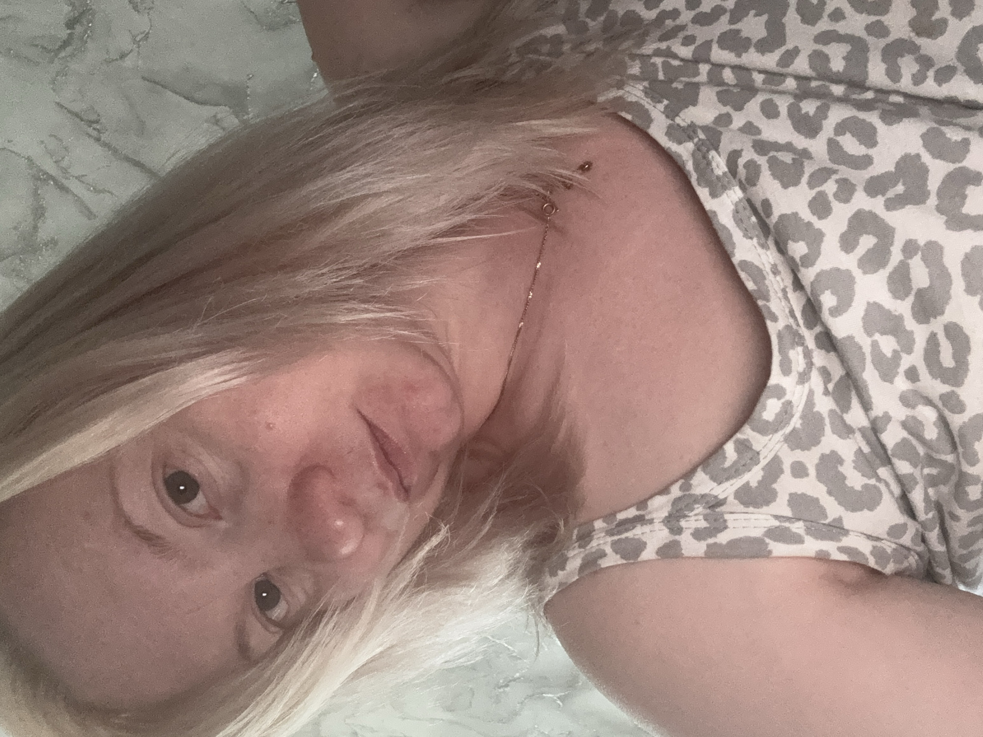 The image is a selfie of a woman with blonde hair. She is wearing a leopard-print tank top. The background appears to be a wall with a textured pattern. The lighting is soft, and the woman's expression is neutral. There is no visible indication of the location being Chaikovsky, Russia.