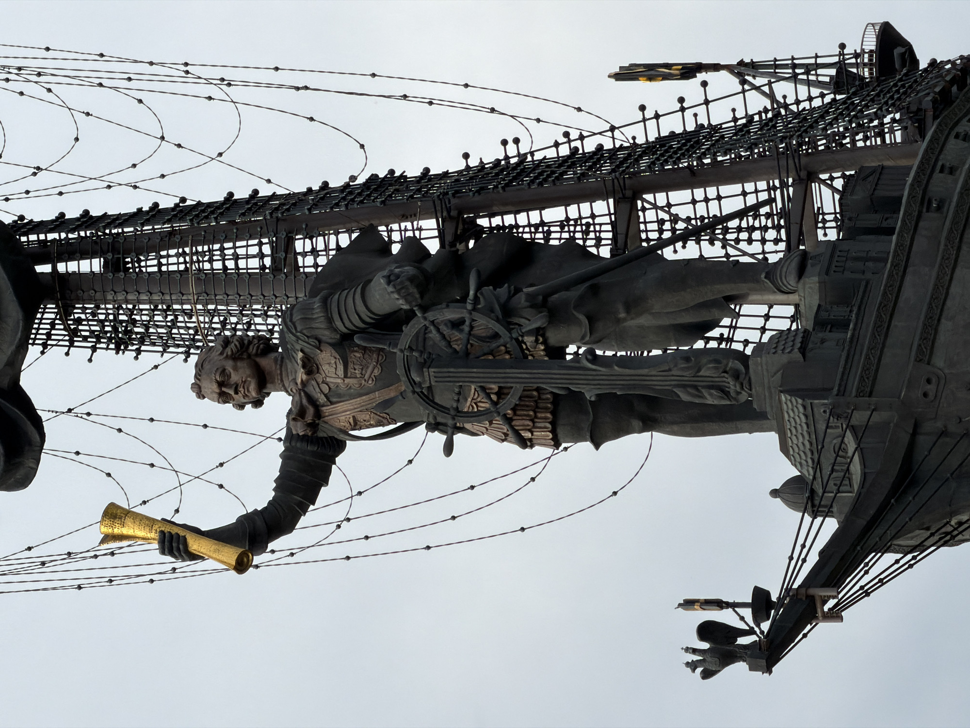 The image depicts a monumental sculpture of Peter the Great in Moscow, Russia. The sculpture, situated against a pale sky, showcases Peter the Great standing at the helm of a ship. He holds a golden scroll in his extended hand. The ship's design is intricate, featuring detailed rigging and decorative elements. The sculpture's material appears to be bronze or a similar dark metal, providing contrast to the sky. The surrounding area is not visible, but the image's upward angle suggests the sculpture's significant height.