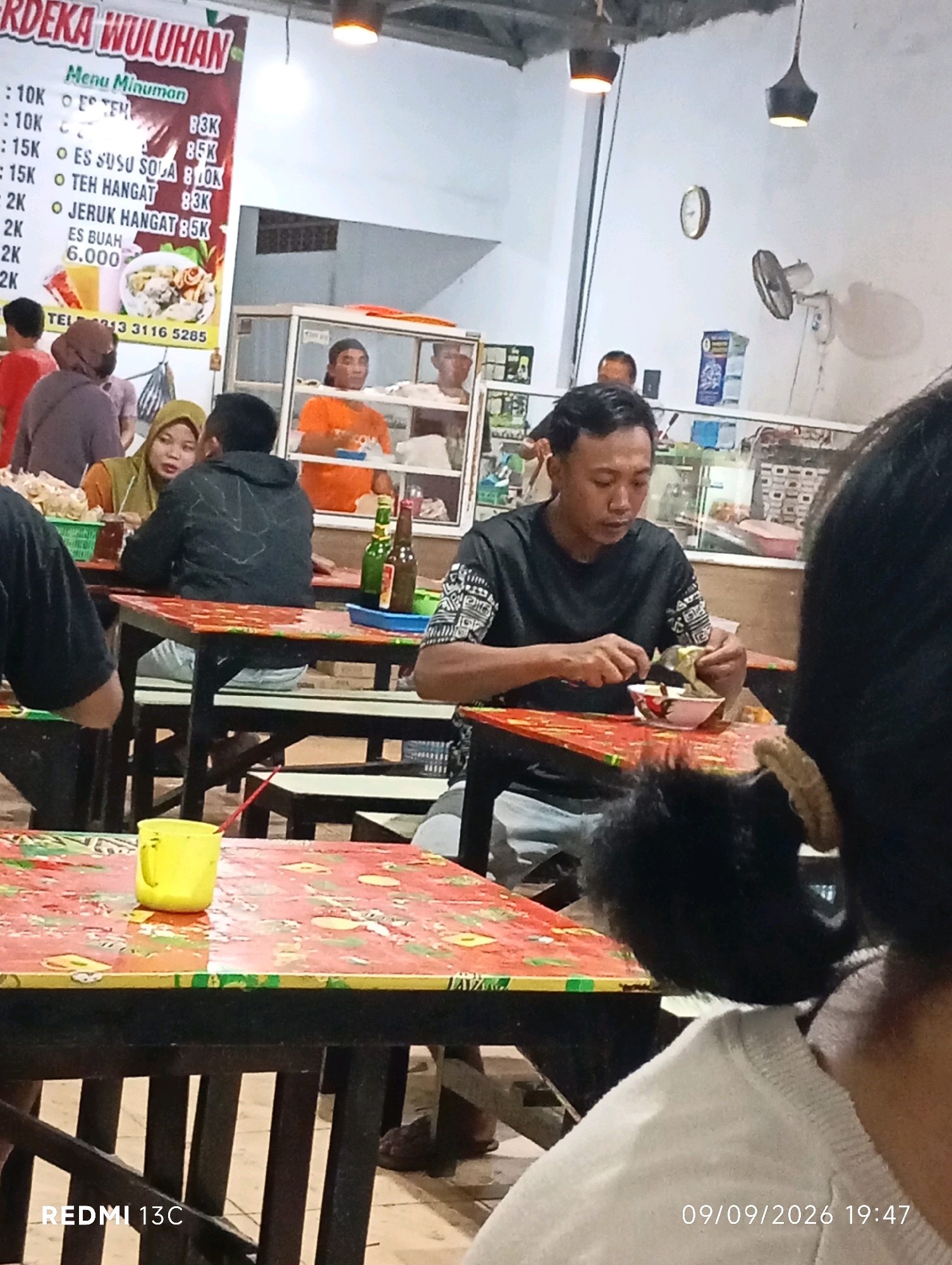 A medium shot captures a bustling indoor food stall in Wuluhan, Indonesia. The scene is filled with patrons seated at tables, suggesting a busy mealtime. In the foreground, a man in a black t-shirt is eating from a bowl with a spoon and fork. To his right, the back of another person's head, with dark hair styled in pigtails, is visible. Yellow cups and red-patterned table cloths are prominent. In the background, a menu board displays prices for various drinks, including "Es Teh," "Es Susu Soda," "Teh Hangat," and "Jeruk Hangat." The stall appears to be open, with a view of the street outside and other customers. The overall impression is one of lively social interaction and casual dining.