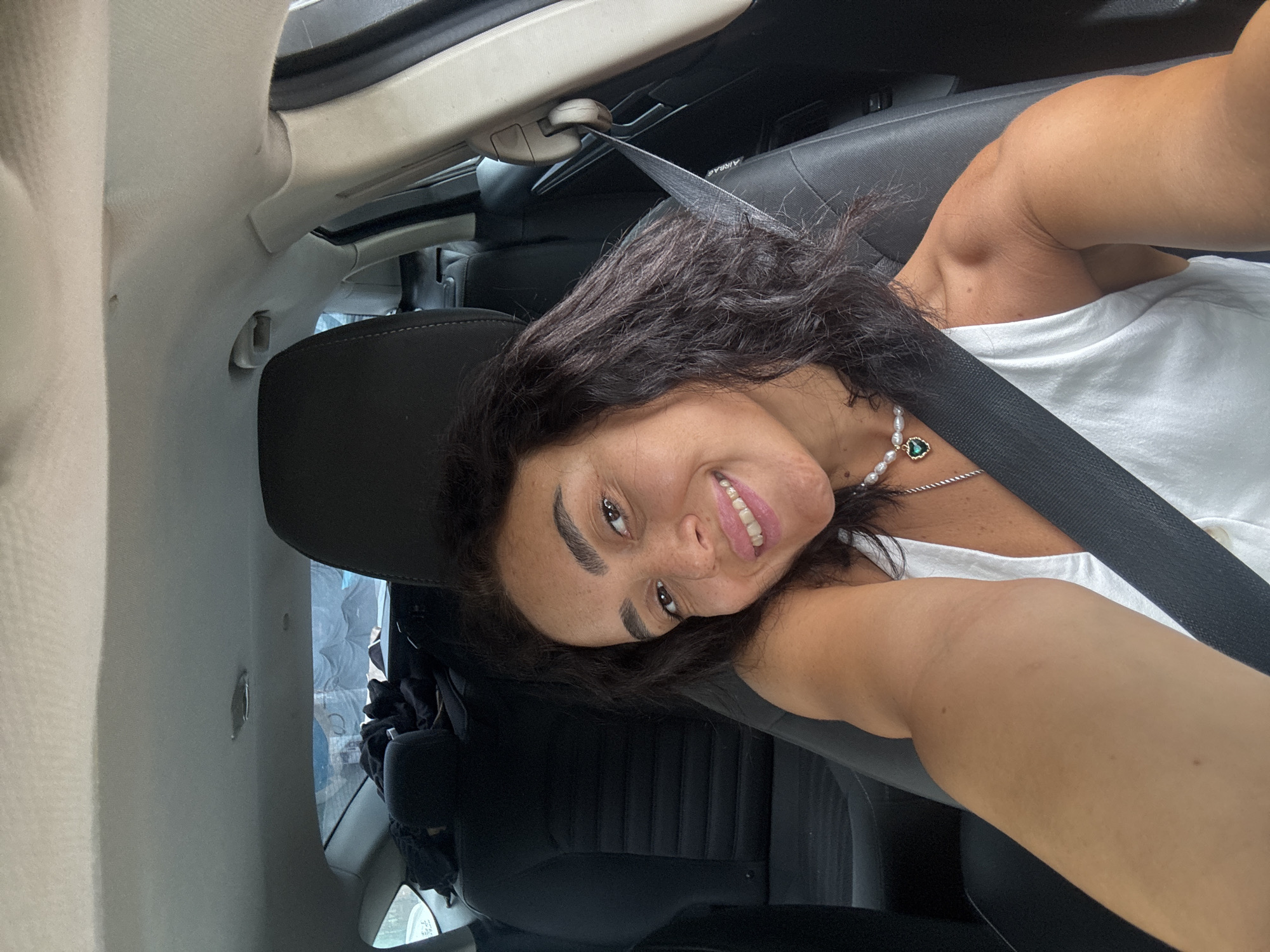 A woman with dark, wavy hair and light brown eyes is taking a selfie in the driver's seat of a car. She is wearing a white tank top and a pearl necklace with a green heart pendant. Her left arm is extended, and she is smiling at the camera. The car's interior is visible, including the black leather seats and the ceiling. The background shows the back seats of the car, which are covered with clothing. The woman is wearing a seatbelt. There is no information available about the specific location in Tbilisi, Georgia, or the time of day.