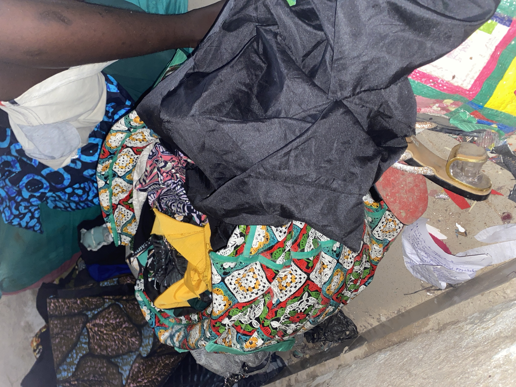 A medium shot captures a jumbled pile of fabrics, predominantly colorful African prints, with a black fabric draped over them. Among the textiles, a yellow garment is visible, along with a white bra and a person's dark-skinned leg and torso. Scattered on the ground are papers, a red shoe sole, and what appears to be a pair of gold-colored sandals. The scene suggests a domestic or personal space, possibly a tailoring area or a storage space for fabrics, located in the city of Jauro Yinu, Nigeria. The lighting is dim, casting shadows and highlighting the textures of the fabrics and the skin.