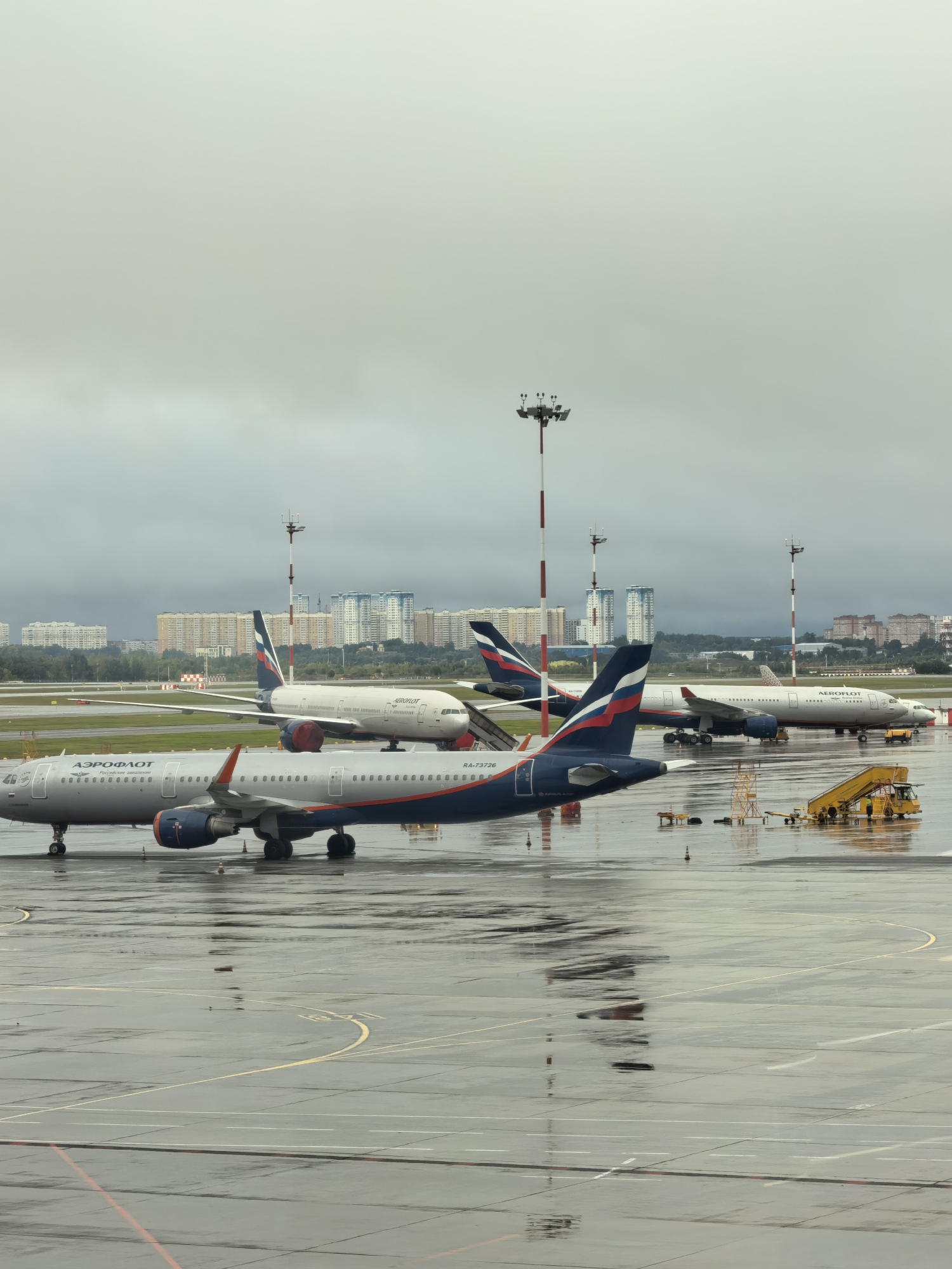 The image depicts an airport tarmac on a cloudy day. Several Aeroflot airplanes are parked on the wet asphalt. In the foreground, an Aeroflot Airbus A321 is visible, along with a yellow passenger stair vehicle positioned near its door. Further back, another Aeroflot airplane, an Airbus A330, is parked, and a third Aeroflot airplane is seen in the distance. The background shows a city skyline with numerous buildings, and tall lighting poles with multiple floodlights are scattered across the tarmac. The ground is wet, indicating recent rain, and reflections of the aircraft and lighting can be seen on the surface.