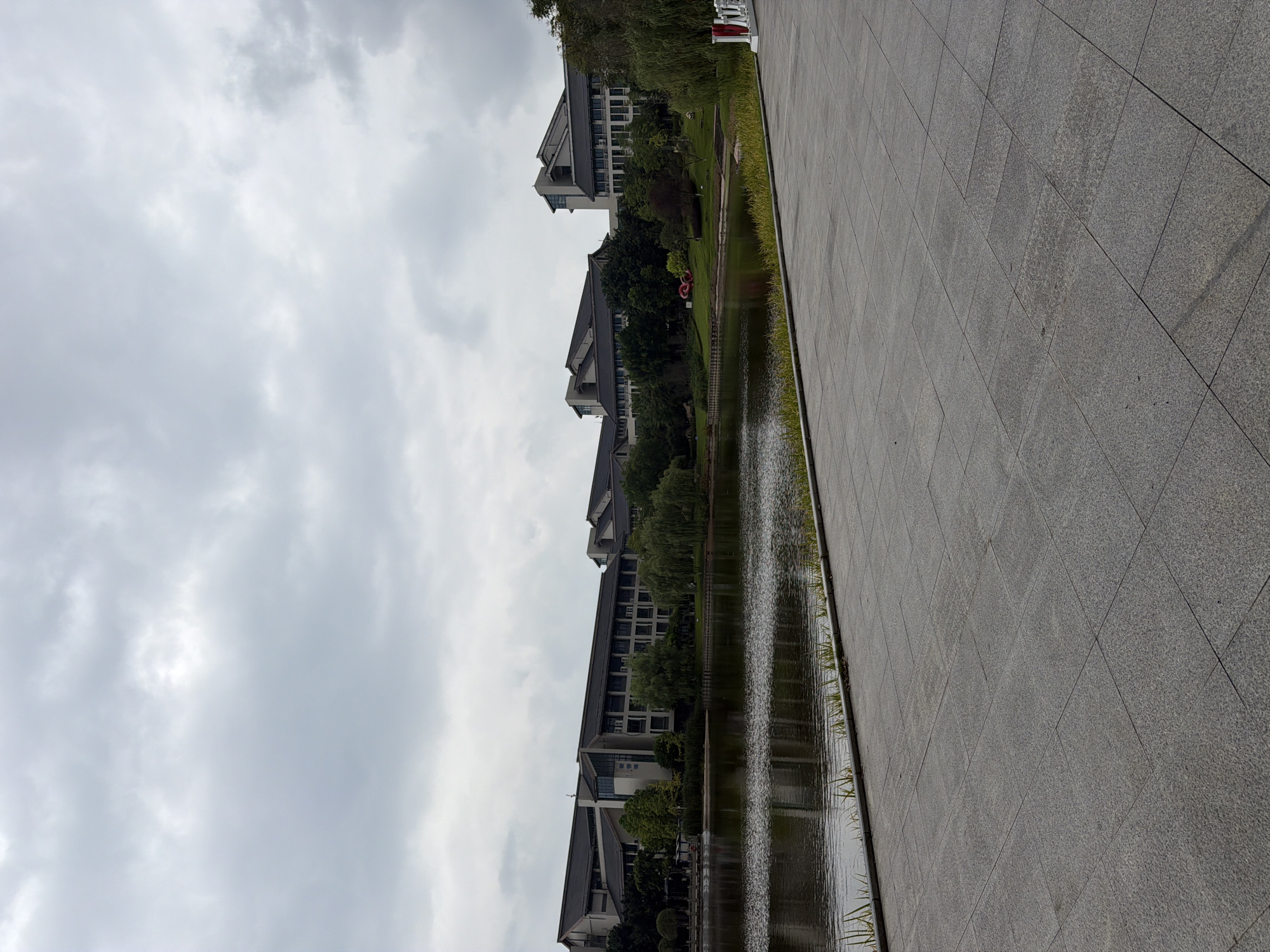 The image depicts a serene lakeside scene with modern buildings in the background, likely part of a university or corporate campus in the city of Jiangjin, China. The foreground is dominated by a paved walkway made of large, gray stone tiles. To the left, a calm body of water reflects the overcast sky and the architecture beyond. Lush green trees, including weeping willows, line the edge of the lake, creating a natural buffer between the water and the buildings. The buildings themselves are multi-story structures with dark, sloped roofs and a contemporary design featuring numerous windows. The sky is heavily clouded, suggesting a dull or rainy day. There are no people visible in the image, and the overall impression is one of quietude and order.