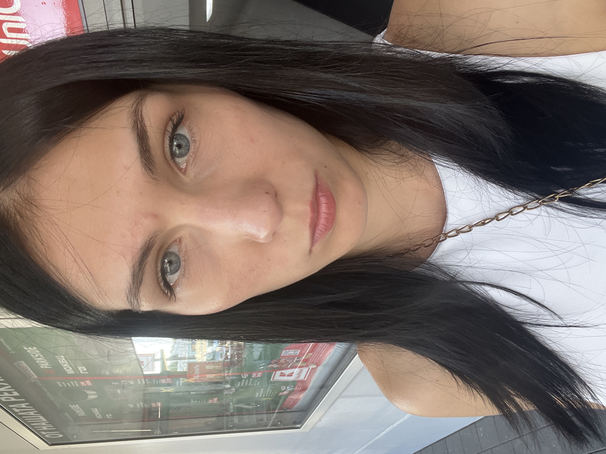 A close-up portrait captures a young woman with long, dark hair and light blue eyes. She is wearing a white tank top with a gold chain necklace. The background appears to be an outdoor setting with a large display board behind her. The display board has text in a foreign language and images of various products. The lighting suggests it is daytime.