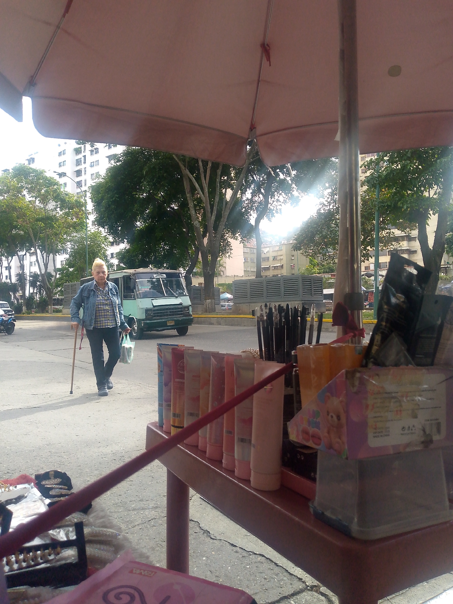The image depicts a street scene in Caracas, Venezuela. An elderly woman with blonde hair, wearing a denim jacket and dark pants, walks across the street using a cane. She is carrying a plastic bag. Behind her, a vintage green bus is parked on the road. In the foreground, an outdoor vendor's stall is partially visible under a large umbrella. The stall displays various items, including cosmetics in tubes, makeup brushes, and packaged goods. The street and surrounding area suggest an urban environment with buildings and trees.