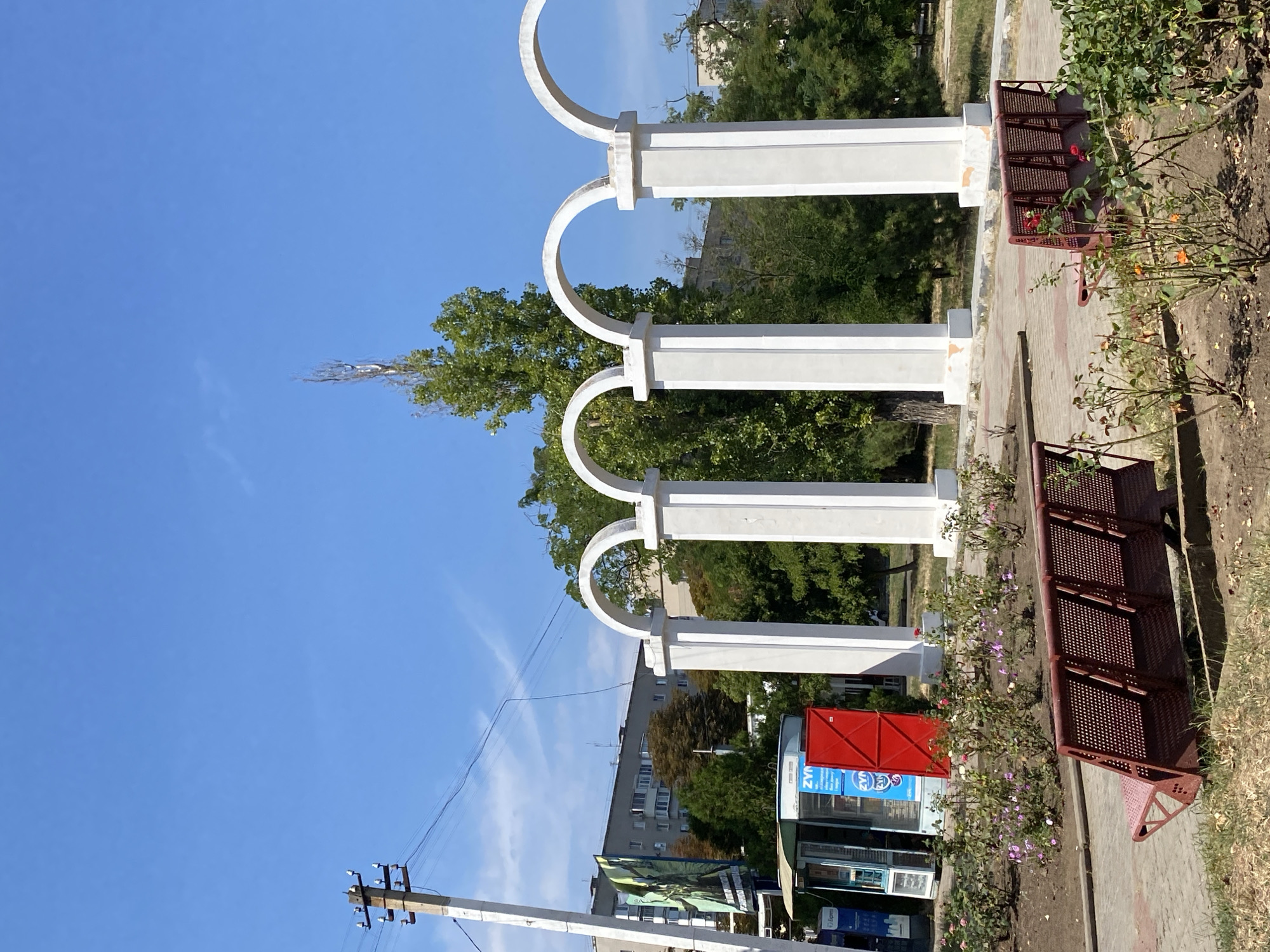 The image depicts an outdoor scene on a sunny day in the city of Chornomorsk, Ukraine. In the foreground, a reddish-brown metal park bench is positioned on a paved walkway. Beyond the bench, a flowerbed with various rose bushes, some in bloom with pink and orange flowers, is visible. To the right, another identical park bench is partially obscured by the rose bushes.
Dominating the midground are several white, arch-topped columns, creating a decorative arcade. Behind the arcade, lush green trees and vegetation are abundant. In the background, a multi-story apartment building with a gray facade stands under a clear blue sky. To the left of the arcade, a small kiosk with a blue and white exterior and a red door is situated. A utility pole with electrical wires is also present on the left side of the image. The overall environment suggests a public park or promenade area.