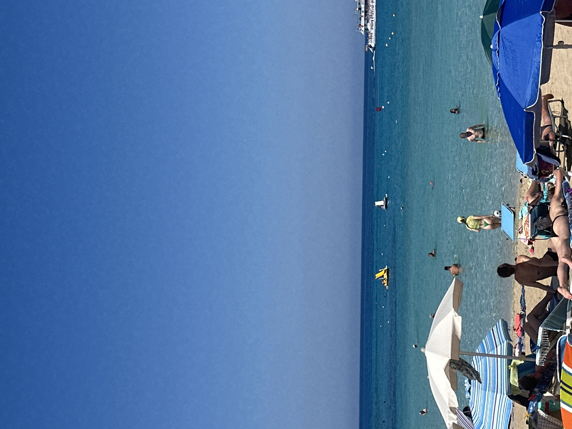 The image depicts a sunny day at a beach in Protaras, Cyprus, with clear blue skies and calm turquoise waters. In the foreground, beach chairs and umbrellas are set up on the sandy shore, with several people relaxing on the chairs. Some are sunbathing, while others are partially obscured by the umbrellas.
In the water, numerous people are swimming and wading. A yellow pedal boat with occupants is visible further out. In the background, a large white boat, likely a cruise ship or ferry, is sailing on the horizon, with "enjoy Coca-Cola" visible on its side. The scene is characteristic of a popular tourist destination, with a lively atmosphere of people enjoying the seaside.