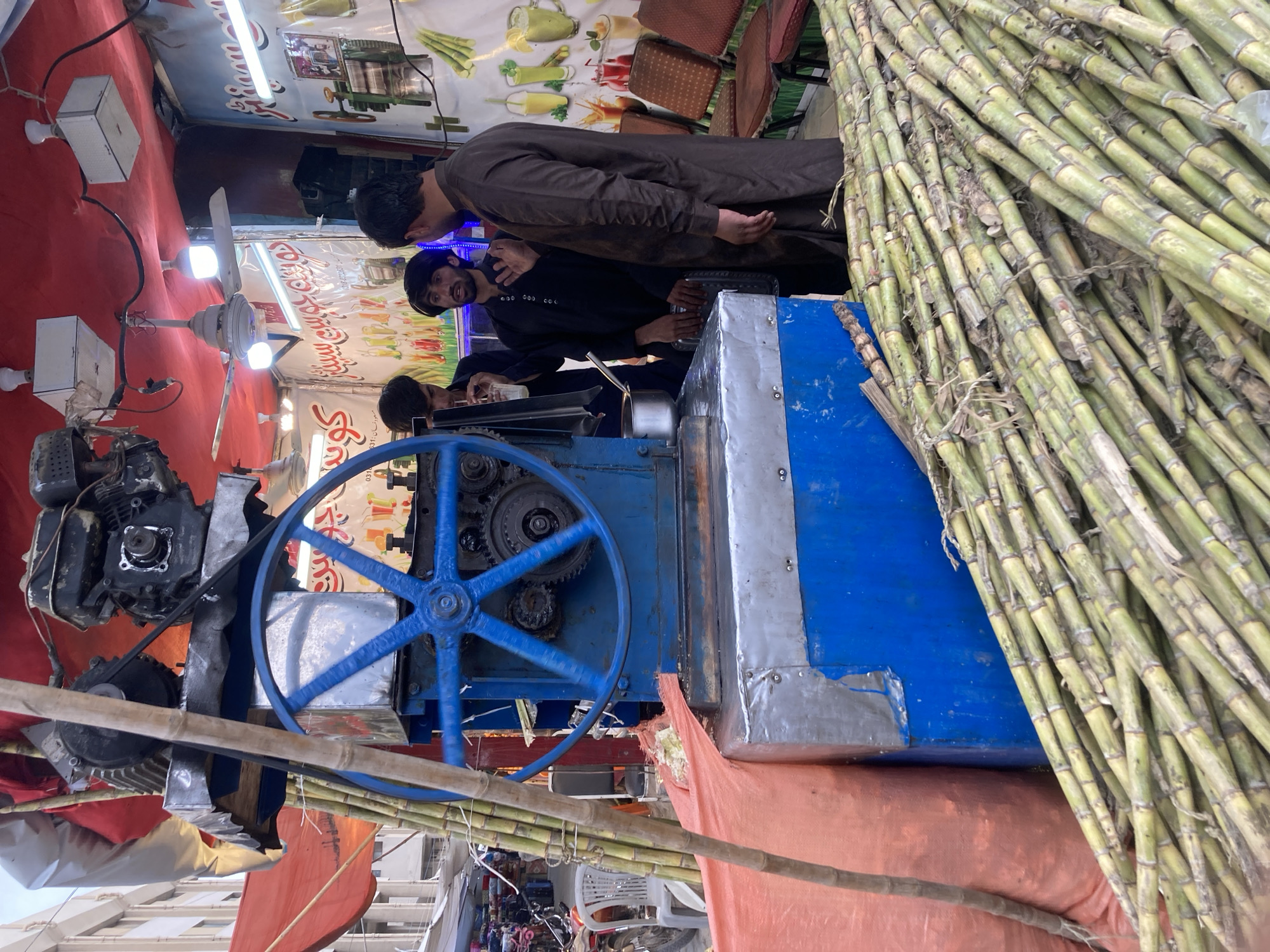 The image shows a sugarcane juice stall in Quetta City Tehsil, Pakistan. There are three men operating the stall. One man in a brown shalwar kameez is standing and watching the other two men who are dressed in black shalwar kameez, who are preparing the sugarcane juice. The sugarcane juice machine is blue and silver, with a large blue wheel on the side. There are piles of sugarcane stalks stacked next to the machine. The stall is decorated with posters of different types of juice. There is a fan hanging from the ceiling of the stall.