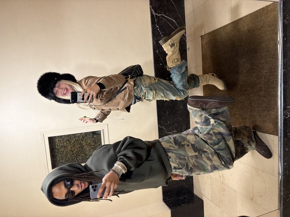 The image is a mirror selfie featuring two individuals, likely taken indoors in what appears to be a hallway or entryway. The entire image is rotated 90 degrees clockwise; describing it as if it were upright:
On the left side of the frame, a person with dark dreadlocks or braids is visible, wearing dark sunglasses and a dark olive green or charcoal grey hooded sweatshirt. They are holding a dark-colored smartphone in their right hand, appearing to take or pose for the selfie. On their left wrist, they wear multiple silver bracelets. Their lower body is dressed in baggy camouflage-patterned cargo pants in shades of green, brown, and black, paired with dark brown low-cut shoes or moccasins.
To the right of the first person, a second individual with long blonde hair is smiling broadly, showing their teeth. They are wearing a large, fluffy black fur hat (ushanka style). Their attire includes a light brown or tan jacket, possibly corduroy, with a collar and visible pockets. They are wearing light blue, distressed or ripped jeans. On their feet are light beige or cream-colored boots with buckle and strap details, extending up the calf. This person is also holding a light-colored smartphone with a distinctive black design on its case in their right hand, seemingly taking the mirror selfie. A black shoulder bag with a thin strap is slung over their shoulder, and a white earbud cord hangs from their ear. Their left hand is slightly open near their waist.
The background consists of a plain, light-colored wall on the left, with a small window or framed opening visible behind the first person. On the right side of the floor, there's a black and white marble or tiled pattern, and further to the right, a dark brown doormat or rug is visible. The lighting suggests an indoor setting, likely well-lit by artificial light or natural light from an unseen source. No discernible text is visible in the image.