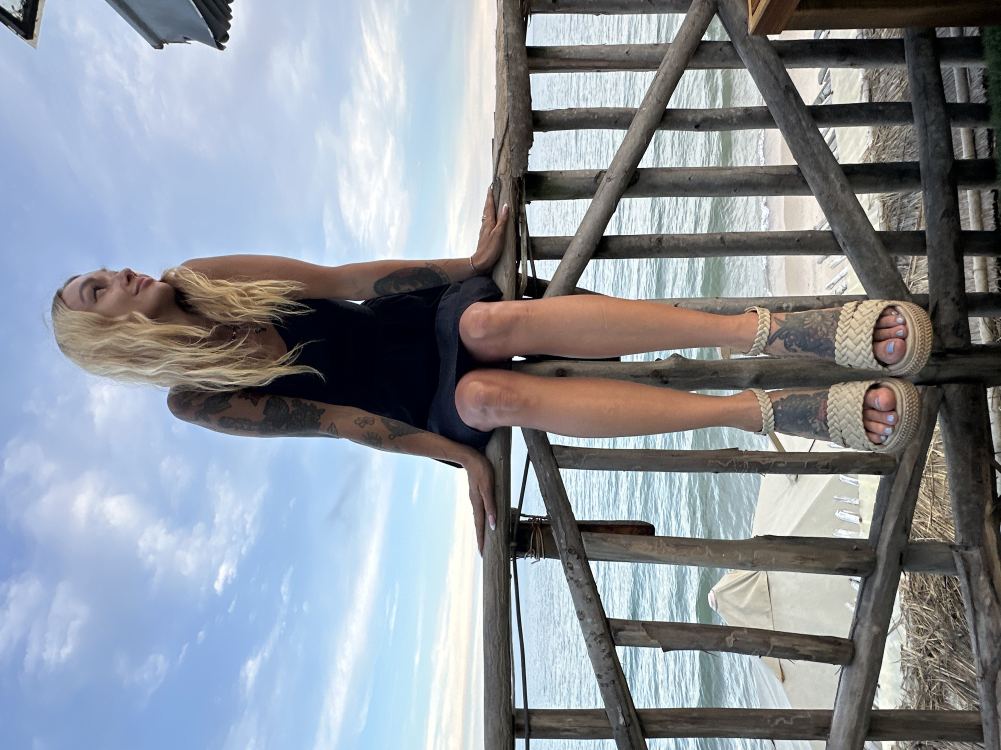 A blonde woman with tattoos on her arms and legs sits on a rustic wooden railing overlooking the ocean. She is wearing a black tank top and a dark skirt, with espadrille wedge sandals. The sky is cloudy, and the ocean is calm with gentle waves. In the background, there are beach umbrellas and some thatched structures. The scene appears to be a coastal resort area in Phu Thuy Ward, Vietnam.