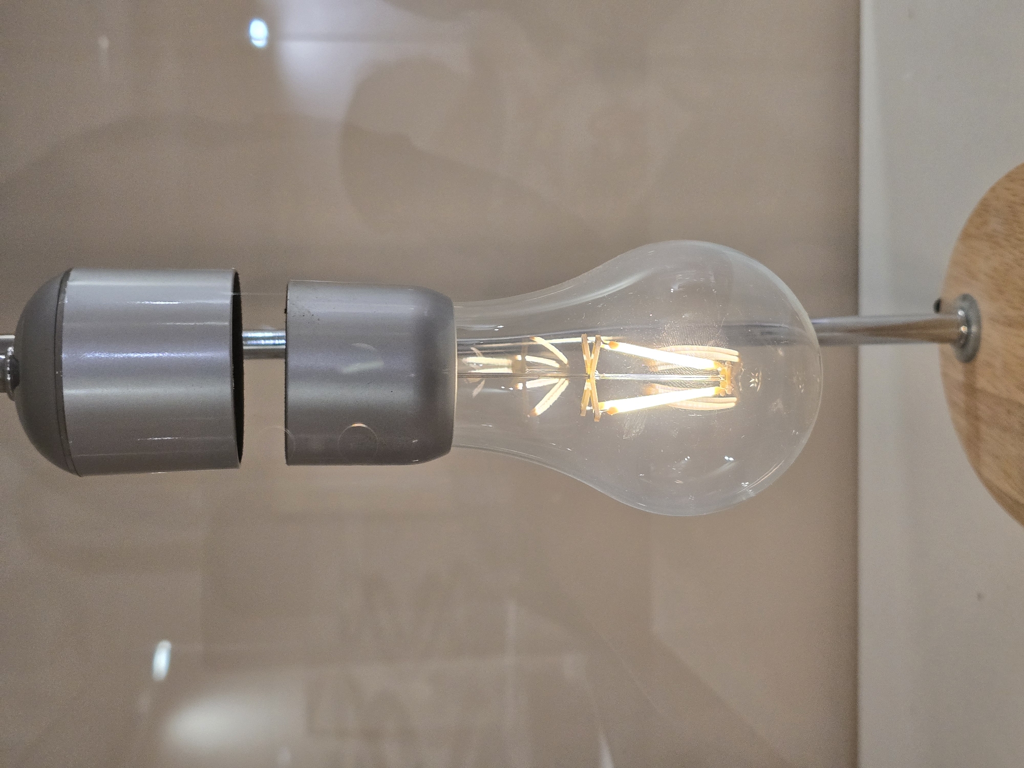 The image depicts a decorative light fixture. It consists of an Edison-style light bulb, encased in a clear glass, affixed to a brushed metal rod. This rod runs through two cylindrical metal sleeves, ending in a circular wooden mounting plate. The background consists of a light beige wall. The location is unspecified.