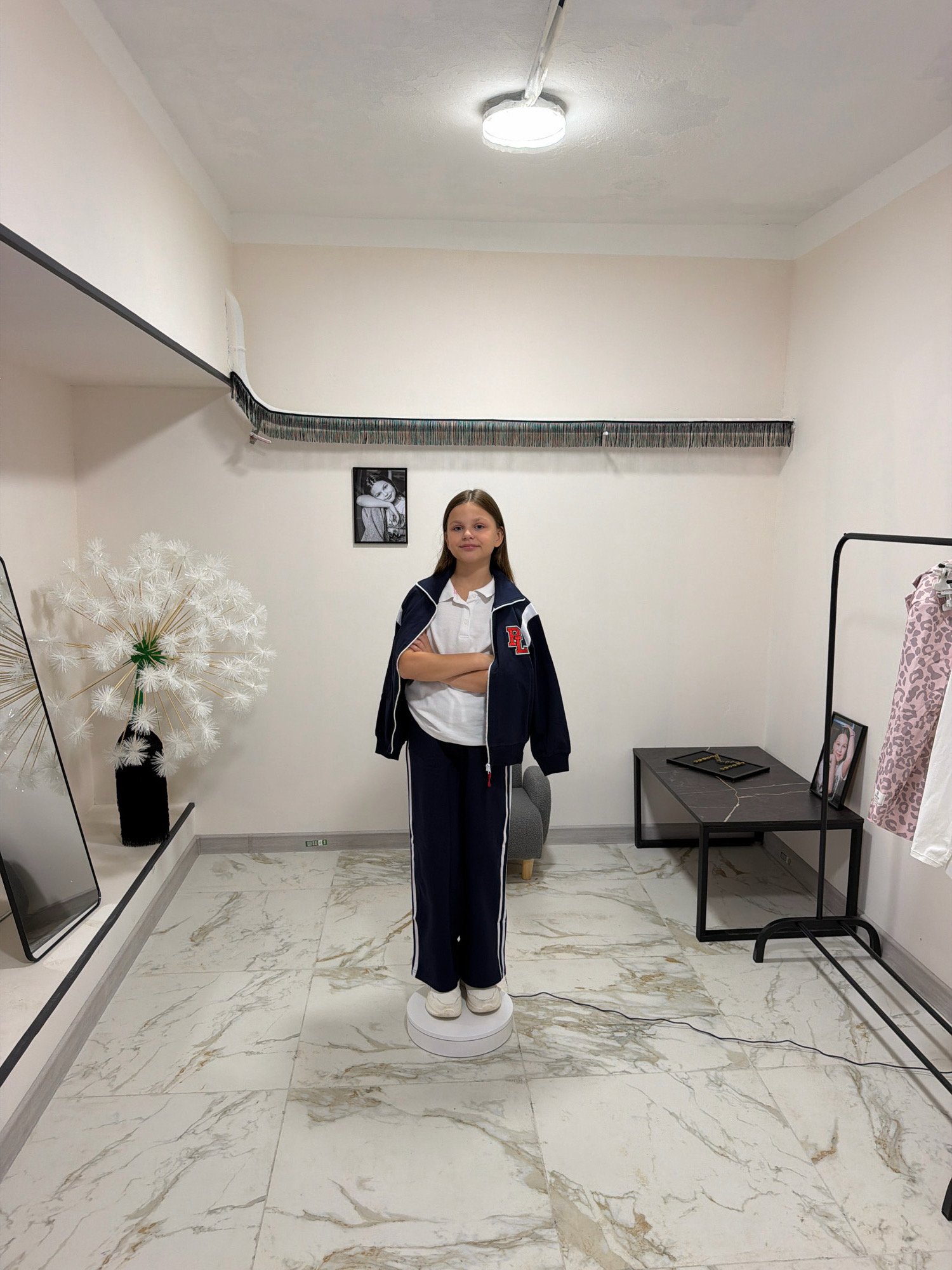 A young female stands on a white rotating platform in the middle of a room with light-colored walls and a marble-patterned floor. She is wearing a white polo shirt, a navy blue jacket with red and white trim, and navy blue pants with a white stripe down the side. Her arms are crossed, and she is looking directly at the camera. Behind her, on the wall, is a framed black and white photograph of a young girl. To her left, a large, white, dandelion-like decoration sits in a black vase. To her right, a black coffee table with a marble top holds a framed item. Further to the right, a clothing rack with several garments, including a pink leopard-print jacket, is visible. The room appears to be a studio or a dressing room, possibly in Khimki, Russia, given the context. A light fixture is visible on the ceiling.