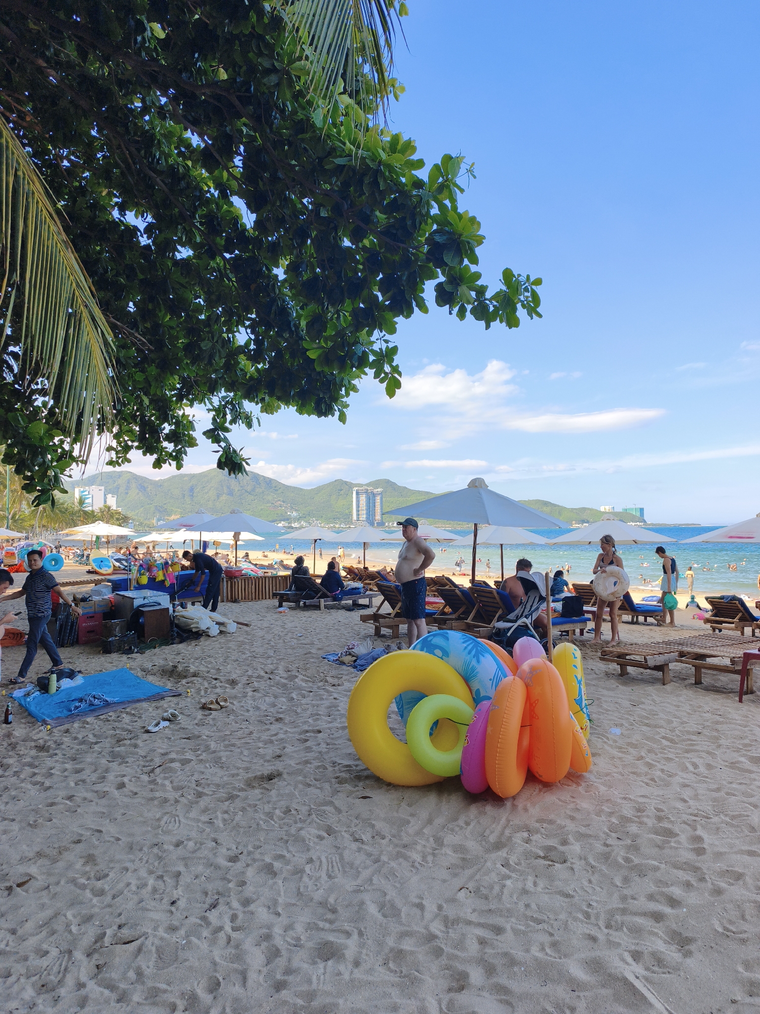 This is a photograph of a crowded beach in Nha Trang, Vietnam, taken on a sunny day. The foreground is dominated by a pile of colorful inflatable rings, a common sight on beaches, indicating recreational activities. To the left, a person is walking across the sand, and there are various items scattered on the beach, possibly belonging to beachgoers or vendors. In the mid-ground, there are numerous beach chairs and umbrellas set up, with people relaxing under them, suggesting a popular tourist spot. Further back, the ocean is visible, with several people swimming or wading in the water. The backdrop features a range of mountains, some with buildings built on them, and the clear blue sky with a few scattered clouds. The overall impression is one of a lively and bustling beach scene in a coastal city.