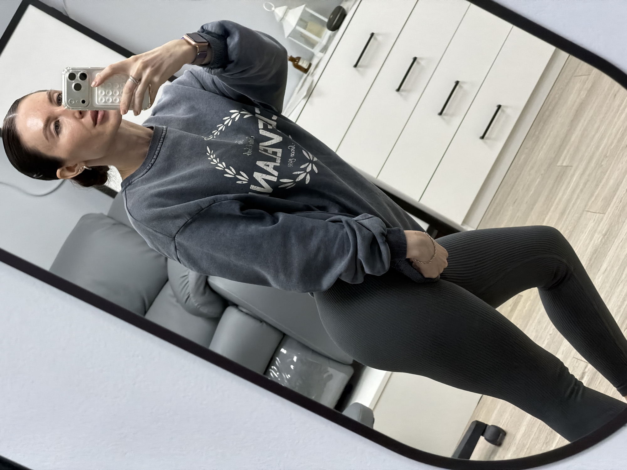 A woman is captured in a full-body mirror selfie, standing indoors in what appears to be a modern home setting in Perm, Russia. She holds a silver smartphone in a clear case up to take the photo, with her gaze directed towards the camera. Her dark hair is neatly pulled back, and she wears a smart watch with a reddish-brown band on her left wrist and a ring on her right hand, along with a thin gold chain bracelet.
She is dressed in casual attire: a dark gray, possibly acid-washed, long-sleeved sweatshirt featuring a distressed white graphic design. The prominent word "MAVEN" is visible in a bold font, encircled by a laurel wreath, with smaller, less legible text below it, possibly "EST. 1993" and "NEW YORK" or "NEW LIFE." She pairs the sweatshirt with form-fitting dark gray or black ribbed leggings.
The background, reflected in the mirror, shows a minimalist interior. To her right, a white dresser or cabinet with black pull handles and a white decorative lantern on top is visible. A light gray upholstered sofa or armchair is partially reflected in the lower-left background. The flooring consists of light-toned wooden planks. The lighting is bright, suggesting daytime. The woman's expression is neutral as she poses for the reflection.