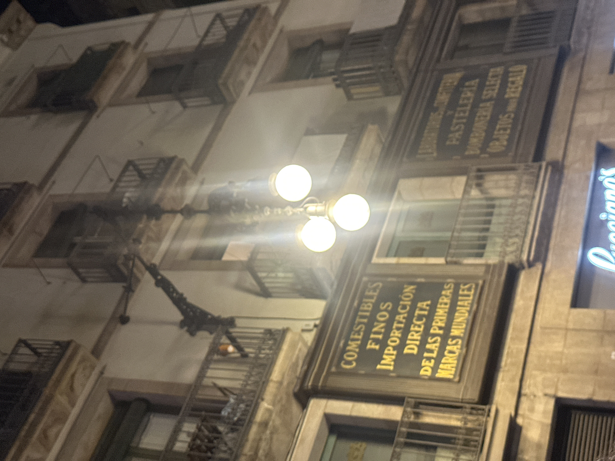 The image captures a nighttime street scene in Barcelona, Spain, focusing on the facade of an old building adorned with illuminated signs and balconies. The architecture is typical of Barcelona's historic districts, featuring ornate balconies with wrought-iron railings and cream-colored stucco walls.
Prominently displayed on the building's facade are two large, dark signs with gold lettering. The sign on the left reads "COMESTIBLES FINOS IMPORTACIÓN DIRECTA DE LAS PRIMERAS MARCAS MUNDIALES," indicating a shop selling fine groceries, direct imports, and world-leading brands. The sign on the right appears to be for a confectionary and pastry shop, possibly stating "LABORATORIOS CONFITERIA PASTELERIA BOMBONERIA SELECTA OBJETOS PARA REGALO," suggesting it sells selected chocolates, candies, and gift items.
A multi-bulb, antique-style street lamp hangs in front of the building, casting a warm glow that illuminates the signs and the lower levels of the facade. The lights create a slightly hazy, atmospheric effect common in nighttime photography. Balconies on multiple levels are visible, some appearing empty, while one on the upper left shows a glimpse of interior furnishings. The ground level of the building is obscured by darkness, but a neon sign with the name "Lucciano's" is partially visible at the very bottom of the frame, suggesting a commercial establishment like an ice cream parlor or restaurant. The overall impression is one of a lively commercial street in Barcelona, characterized by its historic architecture and illuminated storefronts.