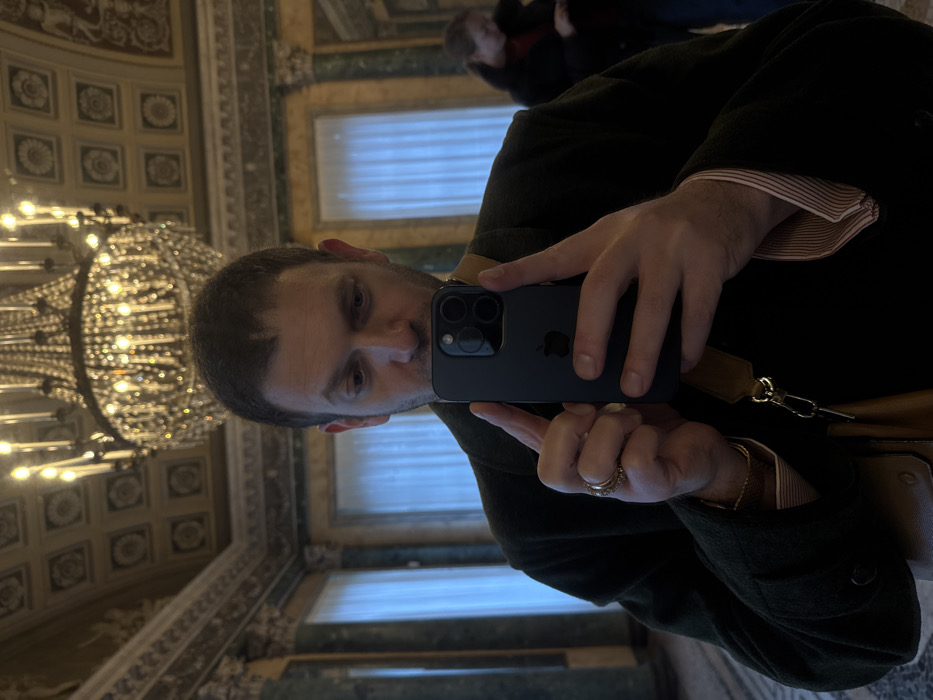 A male subject is captured in a close-up, waist-level selfie reflection, holding a dark-colored smartphone horizontally with both hands, focusing his gaze downwards towards the screen. He has short dark hair and a light complexion, wearing a dark green coat over a light-colored, striped collared shirt with rolled-up cuffs. A gold ring is visible on his left ring finger, and a gold-colored chain bracelet on his left wrist. A brown leather bag strap crosses his upper body.
The background, reflected in the mirror, reveals an opulent interior, characteristic of a grand salon or ballroom in Milan, Italy. The ceiling is elaborately coffered, featuring square panels adorned with detailed gold and cream floral medallions. A large, multi-tiered crystal chandelier with numerous illuminated candle-style bulbs hangs prominently from the center, casting a warm glow and creating bokeh effects. The walls are decorated with architectural elements including fluted pilasters with gilded capitals, ornate cornices, and panels appearing to be made of or finished to resemble dark green marble. Tall windows, covered with translucent white curtains, are visible along the far wall. In the right background, a blurred figure of a second person, dressed in a dark coat, is partially visible, also appearing to look downwards. This grand interior architectural style, particularly the combination of green marble, gilded accents, and detailed coffered ceilings, is consistent with high-end historic venues in Milan, such as **Palazzo Parigi Hotel & Grand Spa Milano**.