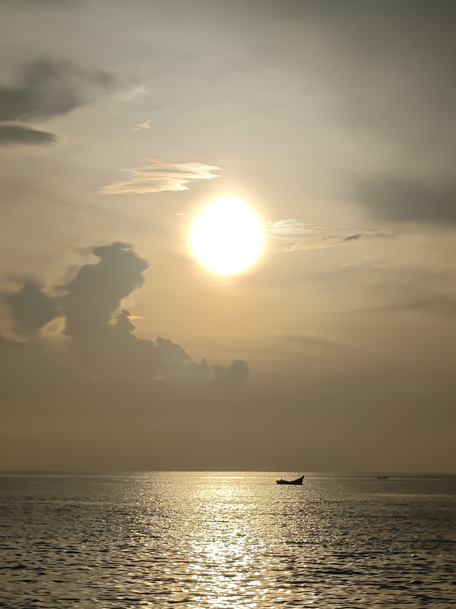 A wide shot captures the golden glow of a sunset over the waters of Lhokseumawe, Indonesia. The sun, a bright orb, dominates the upper center of the frame, casting a shimmering path across the rippling ocean. The sky is a gradient of pale yellow to light gray, with scattered clouds adding texture. A prominent, dark cloud formation resembling a twisted figure stands to the left of the sun, silhouetted against the bright sky. In the middle ground, a small, dark silhouette of a boat drifts on the water, its form barely discernible against the reflective surface. The horizon line is clearly defined, separating the calm sea from the atmospheric sky. The overall impression is one of tranquil beauty at the end of the day.
