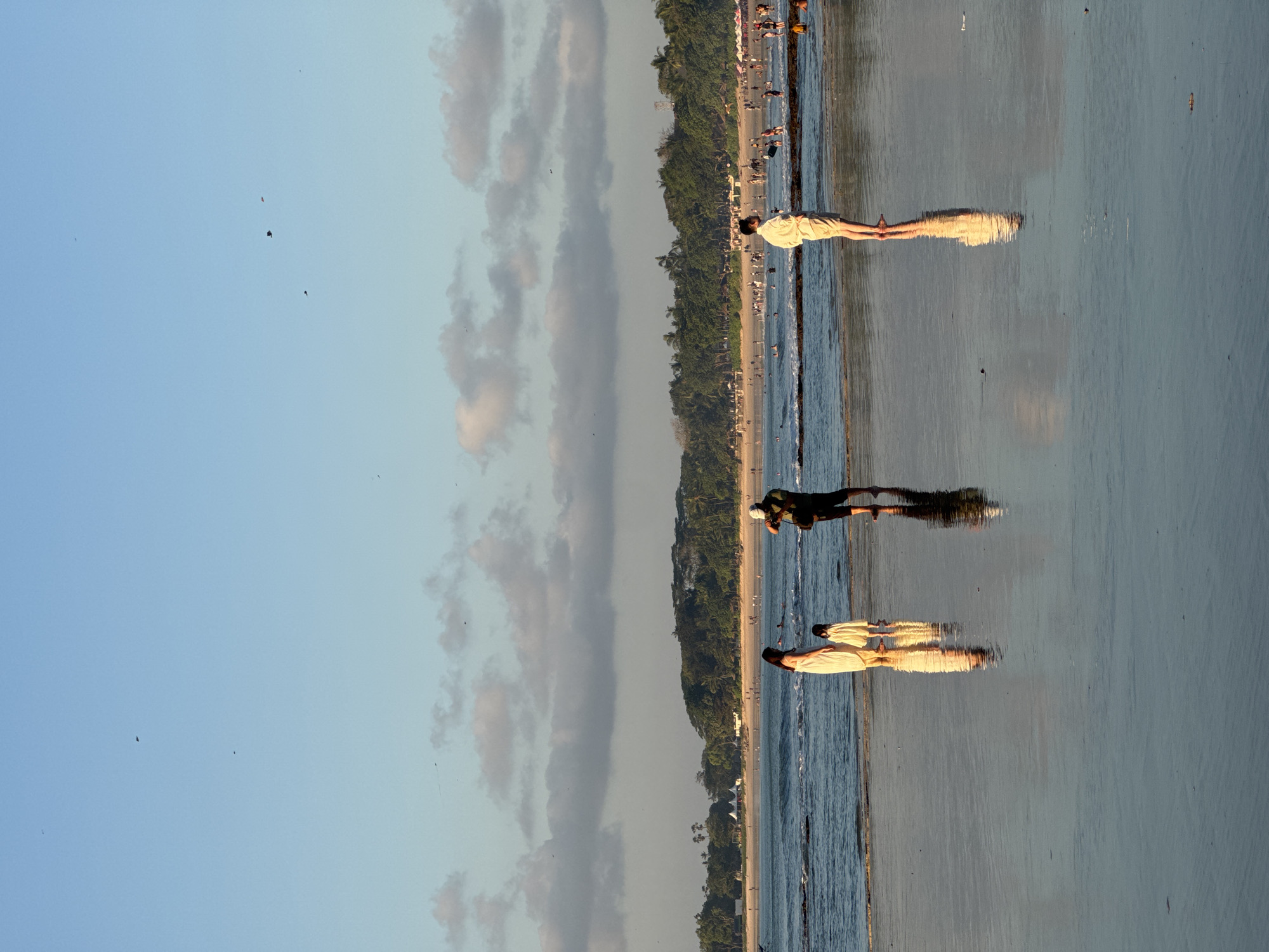 In Kuta Selatan, Indonesia, a wide, sandy beach is illuminated by the warm glow of the setting sun. The tide is low, leaving a vast expanse of wet sand that reflects the sky and the figures on it. In the foreground, three individuals are visible. A woman in a light-colored dress stands with a young child beside her, both facing towards the ocean. To their right, a man wearing a white cap and dark clothing is positioned slightly further back, holding what appears to be a camera and seemingly taking a photograph of the woman and child. Further out on the beach, another person, possibly a man, stands with their back to the camera, wearing a light-colored shirt and shorts. In the middle ground, the ocean stretches out, with gentle waves lapping at the shore. People are scattered throughout the water and along the coastline, enjoying the beach. The background is dominated by a lush green treeline, with buildings and what seem to be white tents nestled within the foliage. The sky is a pale blue, with scattered, elongated clouds near the horizon. Several small, dark shapes, possibly birds or kites, are visible in the sky. The overall scene depicts a serene evening at a popular beach destination.