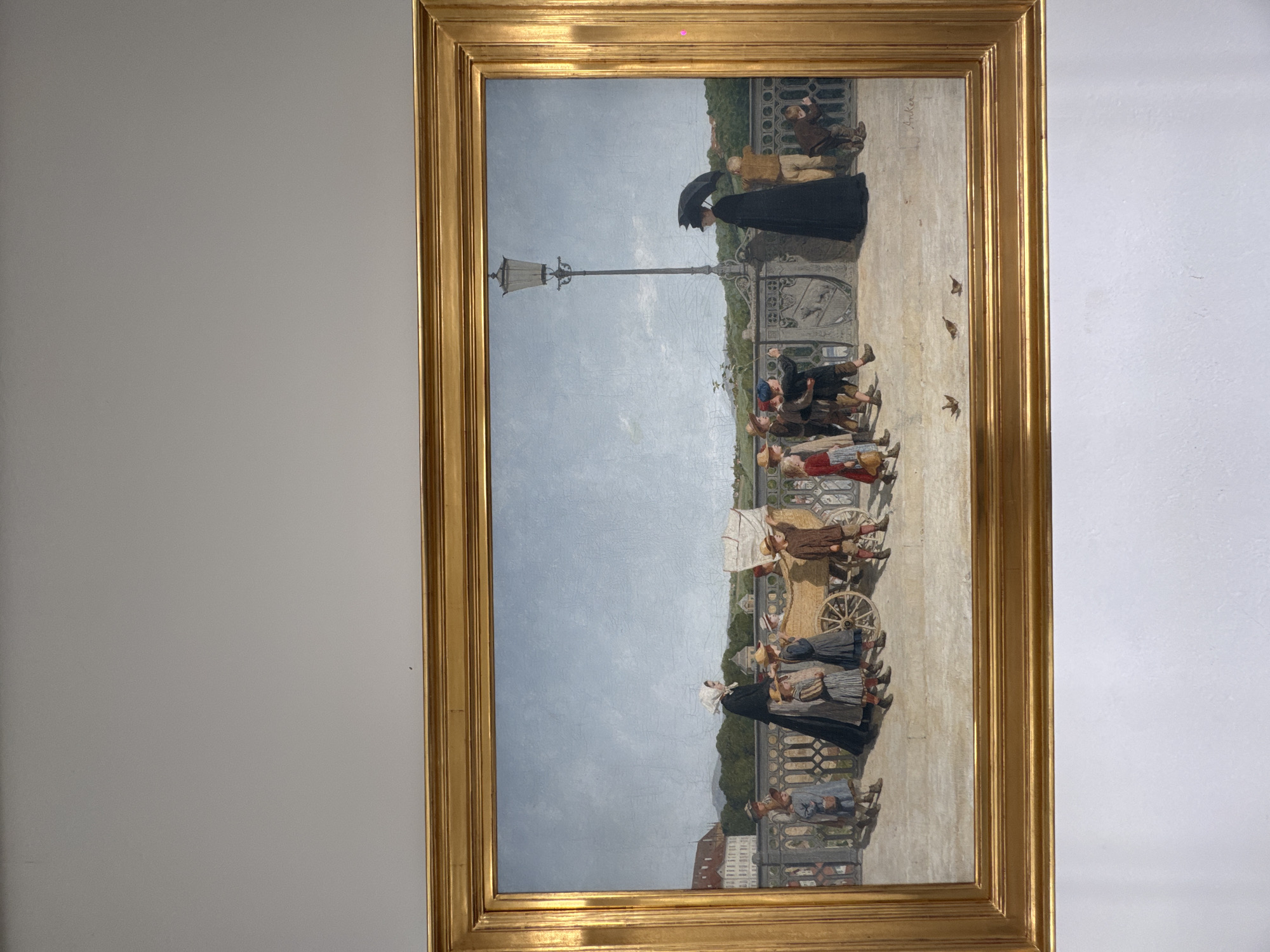 This is a painting in a gold frame, depicting a scene on a bridge or elevated walkway. The viewpoint is from a low angle, looking up at the scene. The sky is overcast, suggesting a daytime setting with perhaps cool or fair weather.
The foreground of the painting shows several groups of people dressed in historical clothing. On the left, a group of children and adults are gathered, some looking towards the viewer, others engaged in conversation. Further back, a woman in a dark, flowing dress holds an umbrella, walking away from the viewer. In the middle ground, a horse-drawn cart with a white canopy is positioned, surrounded by more figures. The people are depicted with detailed clothing and expressions, suggesting a lively or bustling atmosphere.
In the background, a bridge railing with an ornate pattern separates the walkway from a landscape beyond. This landscape includes trees and what appears to be a city with buildings, possibly in Bern, Switzerland, given the location context. A lamppost stands prominently on the walkway, adding to the urban feel of the scene. A few birds are visible flying in the sky.
The painting captures a moment in time, possibly a public gathering or a stroll along the promenade. The overall mood seems relatively calm, though the interaction between the figures suggests a sense of everyday life and social activity. There is some visible cracking in the paint surface, indicative of age. No discernible text is present in the visible portion of the painting.