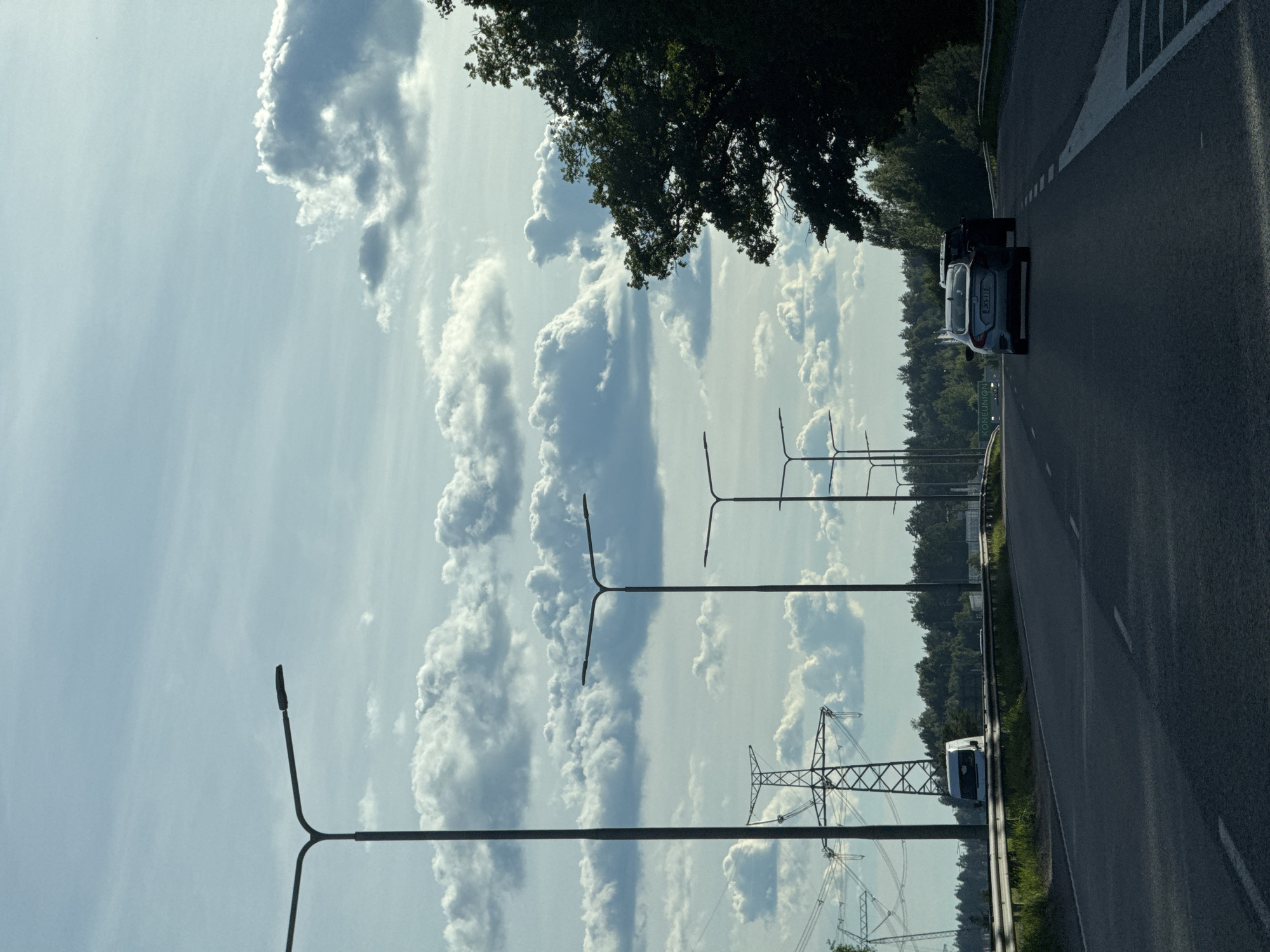 The image depicts a road scene on a cloudy day. In the foreground, a dark asphalt road stretches into the distance, marked with white lane lines. Several streetlights are visible along the side of the road. In the middle ground, a white van is parked on the side of the road, with tall electrical towers and power lines behind it. Further down the road, two cars are driving away from the camera, with trees and foliage lining the background. The sky is filled with large, white, puffy clouds against a light blue backdrop. The overall impression is of a highway or a major road in a semi-urban or rural setting.