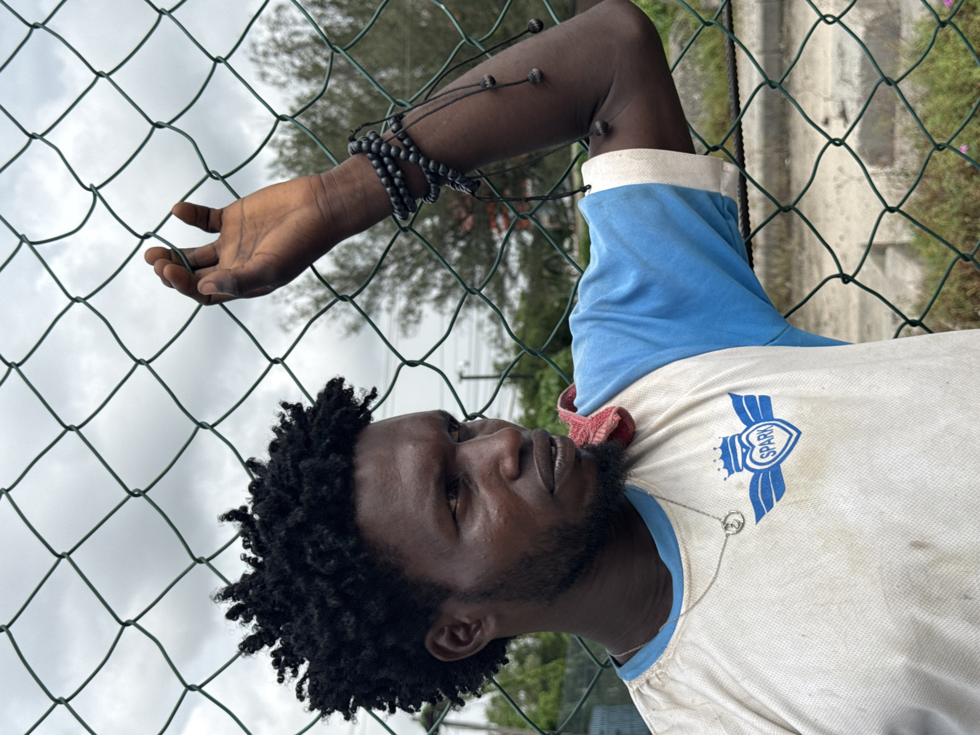 This portrait-style image, taken in Lekki, Nigeria, features a Black man from the chest up, positioned against a green wire mesh fence. The man has dark, curly hair and a short beard. He is wearing a white short-sleeved collared shirt with blue sleeves and collar. On the chest of his shirt, a blue logo depicts a heart with the word "SPARK" inside, flanked by wings, and topped with a crown. A silver necklace with a circular pendant is visible around his neck.
His left arm is extended, with his hand resting gently on the wire mesh fence, appearing to reach or touch it lightly. His right arm, partially visible, is adorned with several dark beaded bracelets. He is looking upwards and to his left, with a contemplative or stoic expression.
The background consists of an overcast, cloudy sky, suggesting daytime. Through the fence, glimpses of green foliage and a concrete structure are visible. The man's shirt shows some signs of wear and minor stains. The prominent fence in the foreground creates a sense of separation or looking through a barrier.