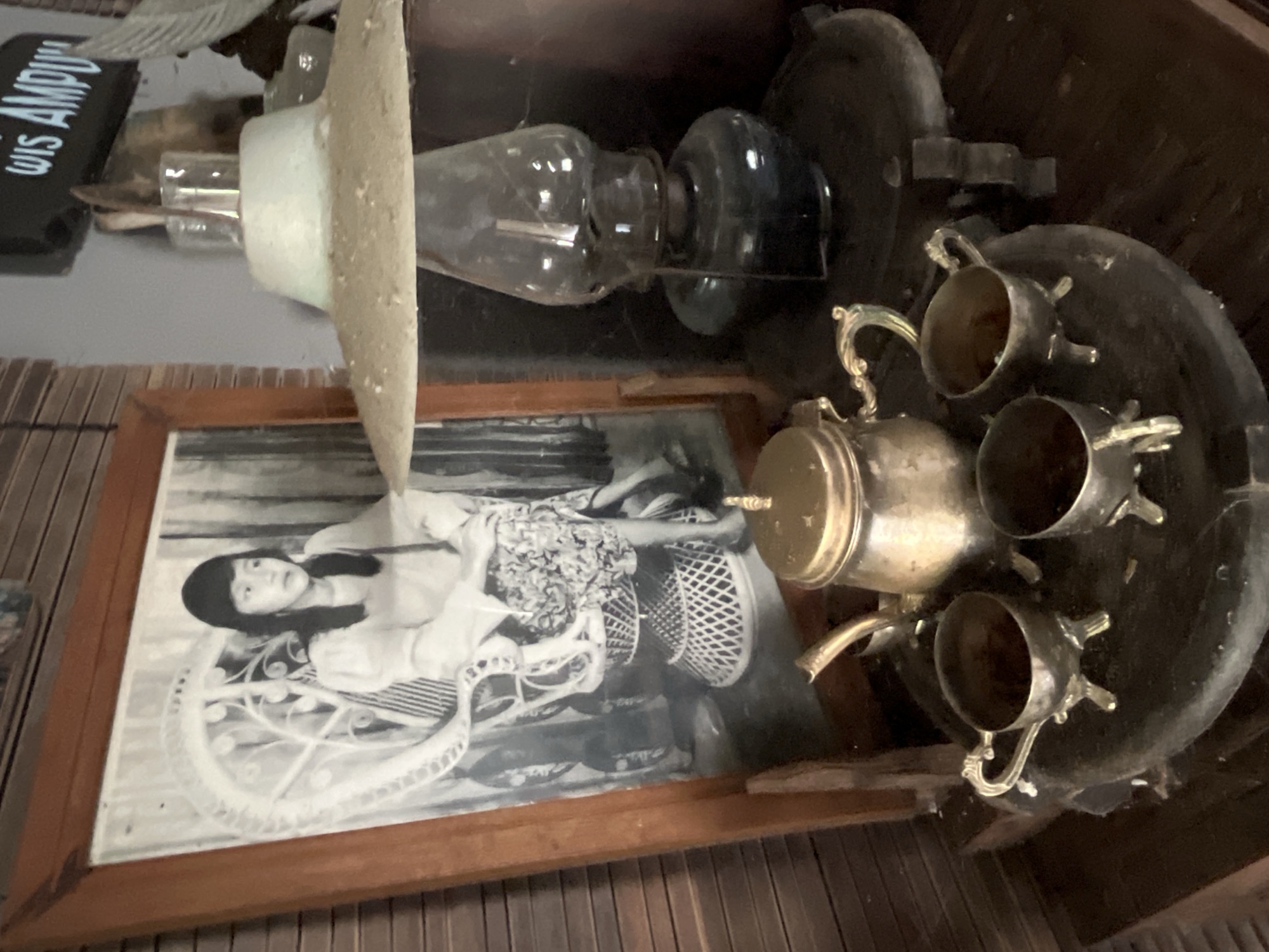 A vintage still life arrangement is captured from a medium shot, foreground to background. The scene is set indoors, with a dark wooden slatted wall serving as the backdrop. On the left, a framed black and white photograph of a young woman seated in a wicker chair is prominently displayed. The woman has bangs and is wearing a puffed-sleeve top and a patterned skirt. To the right of the photograph, a kerosene lamp with a weathered, conical shade sits atop a dark, circular pedestal. The lamp has a glass reservoir and a dark metal base. In the immediate foreground, a small, ornate gold teapot with a curved spout and a lid is placed on a round, dark surface. Three matching gold teacups, each with delicate handles and resting on three small legs, are arranged around the teapot. The overall atmosphere is one of nostalgia and antique decor, possibly within a traditional Indonesian home in Tambakreja, Indonesia, given the context of the location.