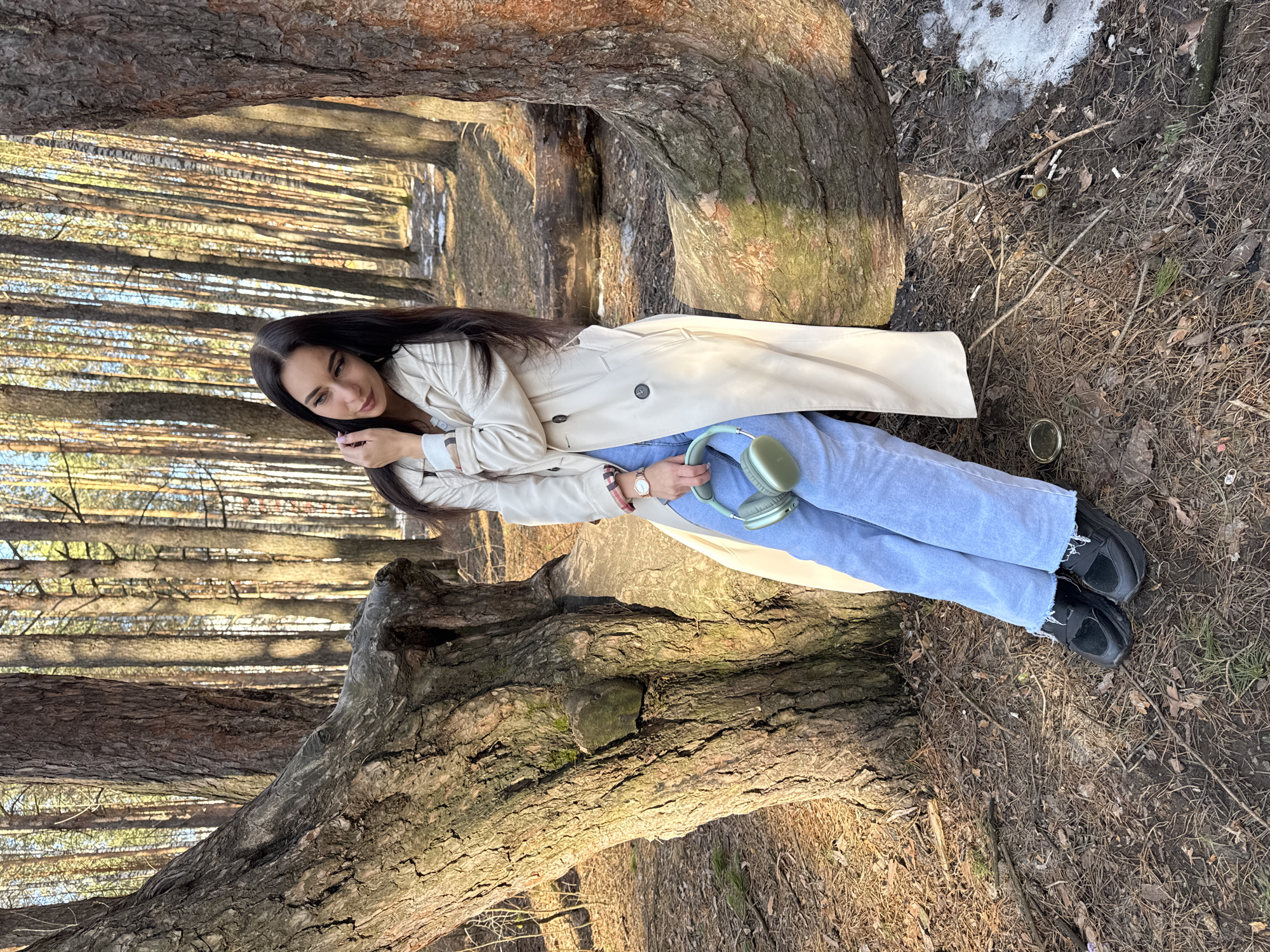 A woman is reclining within the confines of a large, fallen tree in a forest. She is wearing light blue jeans, black shoes, and a beige trench coat. She is holding mint-green headphones. The location is likely in a natural environment, possibly near Chaikovsky, Russia, though the specific city cannot be verified from the image alone. The ground surrounding the tree is covered in leaf litter and debris. A small patch of snow is visible in the background. The forest contains numerous tall trees. There are discarded metal cans and cigarette butts near the tree on the ground.