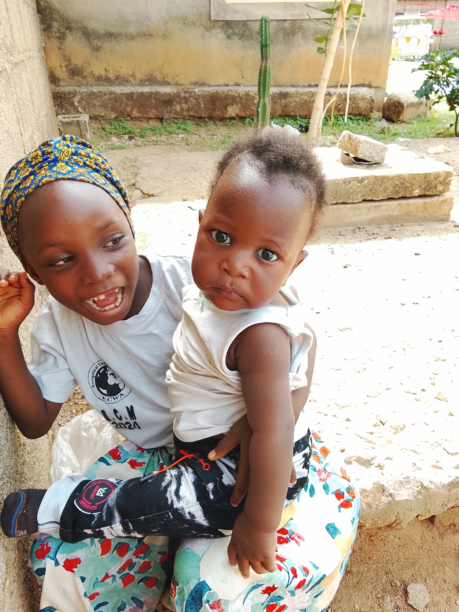 Two Black African girls sit outside a building. The older girl wears a blue and yellow head wrap, a white t-shirt with the logo "Evangelical Church Winning All (ECWA) L.C.M 2024", black pants with white designs and blue socks. She is missing a front tooth and has her arm raised. The younger girl is being held by the older girl, has a fair complexion and has on a white shirt and black and white pants. The seated girl is sitting on a floral print skirt. There is a cement building, a cactus, a small plant, and a rock visible. This image was taken in Akwanga, Nigeria.