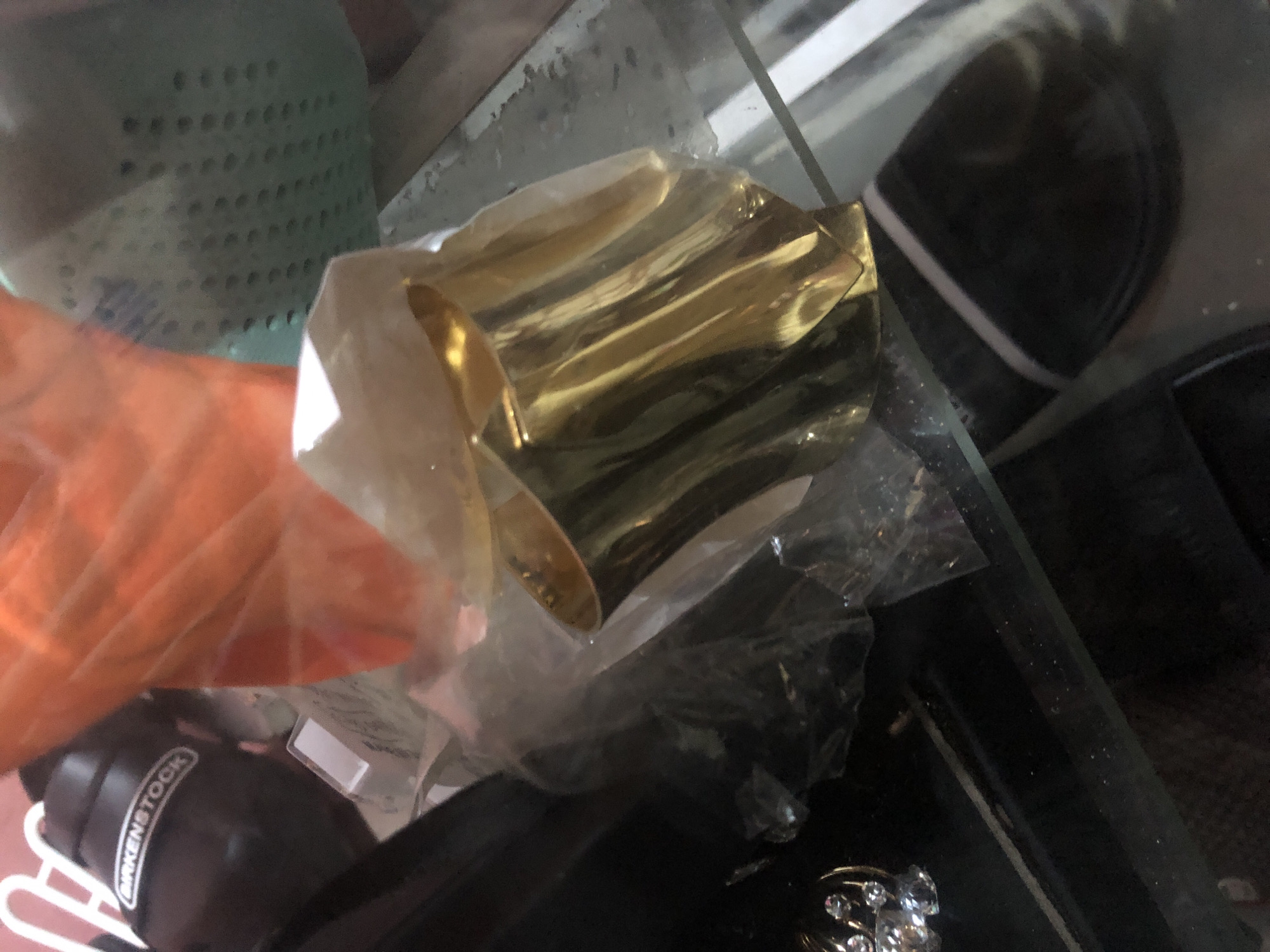 The image shows a close-up view of several items displayed on a glass surface, possibly in a market stall or shop. In the foreground, a gold-colored, wide cuff bracelet with a textured, wavy surface is the prominent object. It is partially covered by clear plastic packaging. To the left of the bracelet, a dark cylindrical object with a white band and text that appears to read "BIRKENSTOCK" is visible. Behind these items, there are out-of-focus elements including what looks like orange plastic sheeting and a mint green textured object. The overall environment appears to be indoors, with artificial lighting casting reflections on the glass surface. The scene does not provide any specific geographical markers that would indicate its location within Ado, Nigeria.