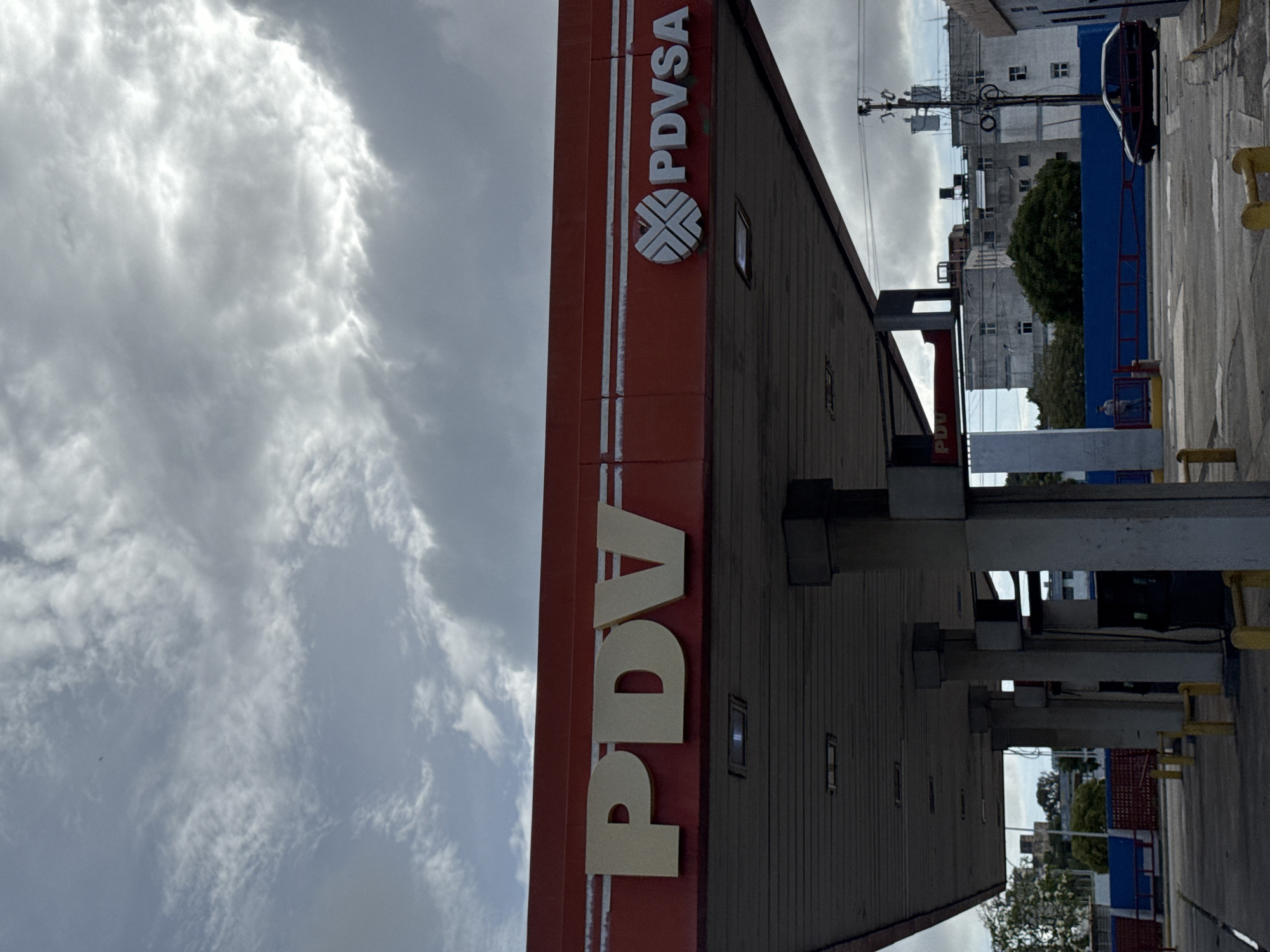 The image shows a PDVSA gas station in Porlamar, Venezuela. The station features a red canopy with large, white "PDV" lettering and the PDVSA logo. Beneath the canopy, multiple gas pumps are visible, supported by concrete pillars. In the background, several multi-story buildings and trees are present under a cloudy sky. A dark-colored car is parked near one of the pumps, and a person stands by a fence in the middle ground. The overall scene depicts a typical urban environment on what appears to be an overcast day.