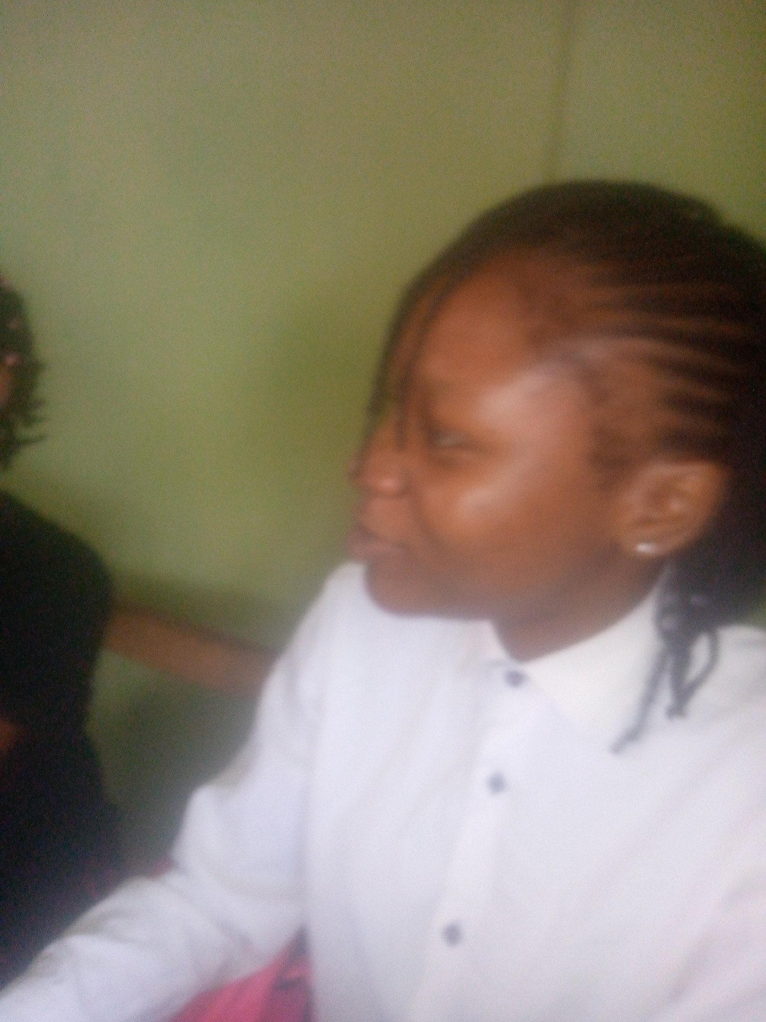 The image shows a woman in a white collared shirt, viewed from the side. Her hair is styled in cornrows with some strands hanging down. She is wearing an earring in her left ear. Part of another person is visible in the left of the frame. The background appears to be a plain, light green wall. The location in Ikorodu, Nigeria is not discernible from the visual information provided in the image.