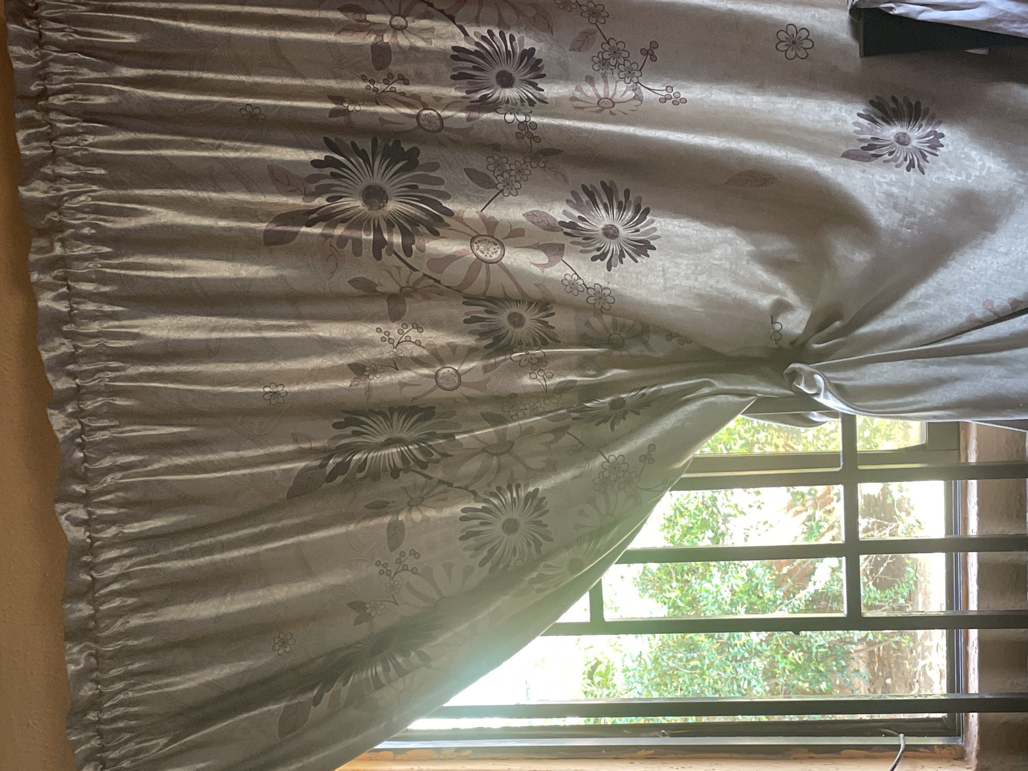 The image shows a light-colored curtain with a floral pattern, partially drawn to reveal a window. Through the window, lush green foliage is visible, suggesting an outdoor environment. The window is covered by a metal grille. The curtain is tied back on the left side with a fabric band. The lighting suggests it is daytime, with sunlight streaming in from the window. The overall scene depicts a room interior looking out onto a garden or natural landscape. This setting could be a home or an accommodation in Kakau, Nigeria.