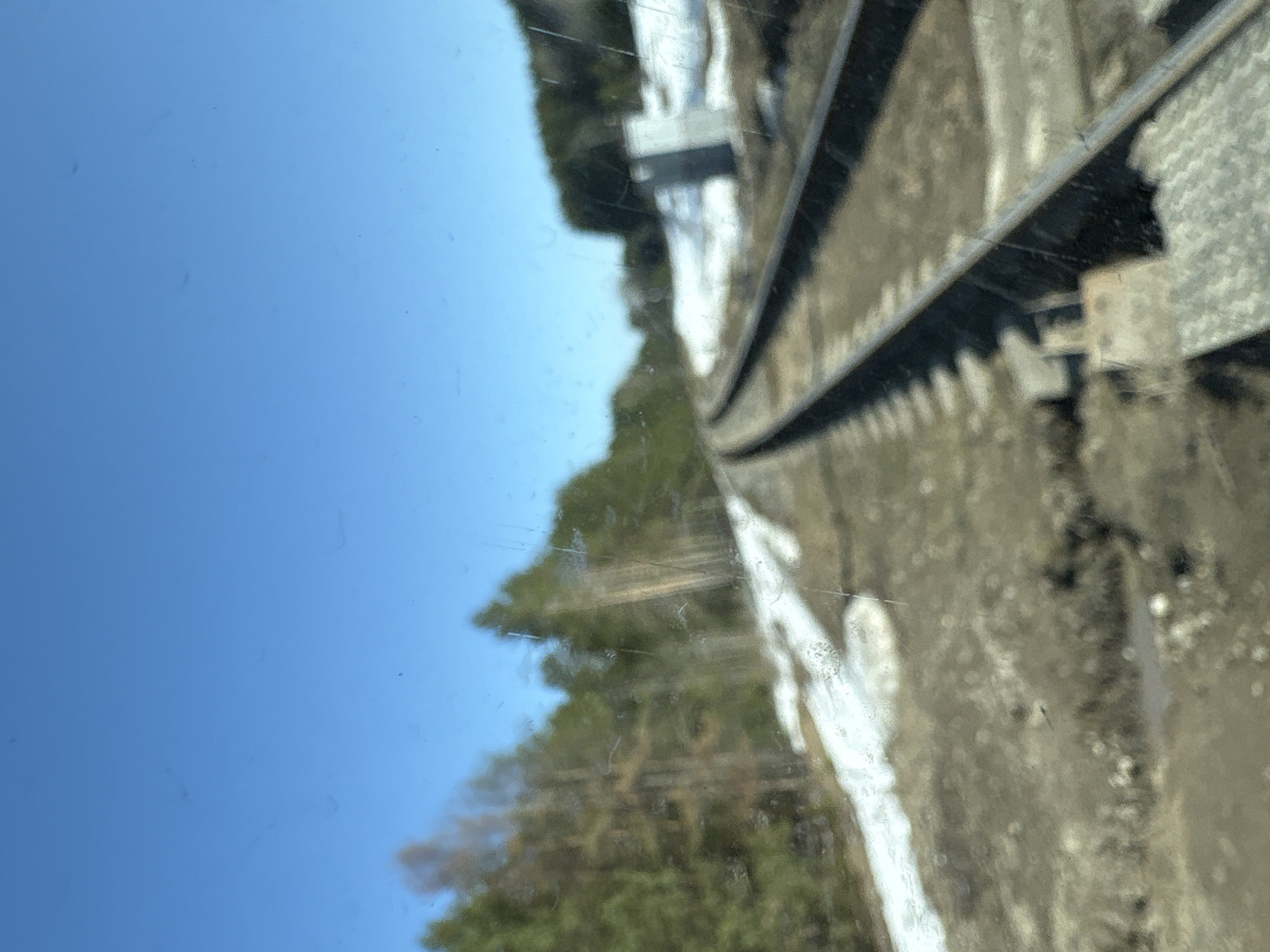 The image shows a view from a window, likely of a train or building overlooking railway tracks. The sky is blue and clear, visible on the left side of the frame. Trees are partially visible through the glass, suggesting a rural or semi-rural setting. The railway tracks are the dominant feature, with visible rails and ballast. The presence of possible snow or ice on the ground along the tracks. There is no evidence to determine the location is in Chaikovsky, Russia. There are no people visible in the image.