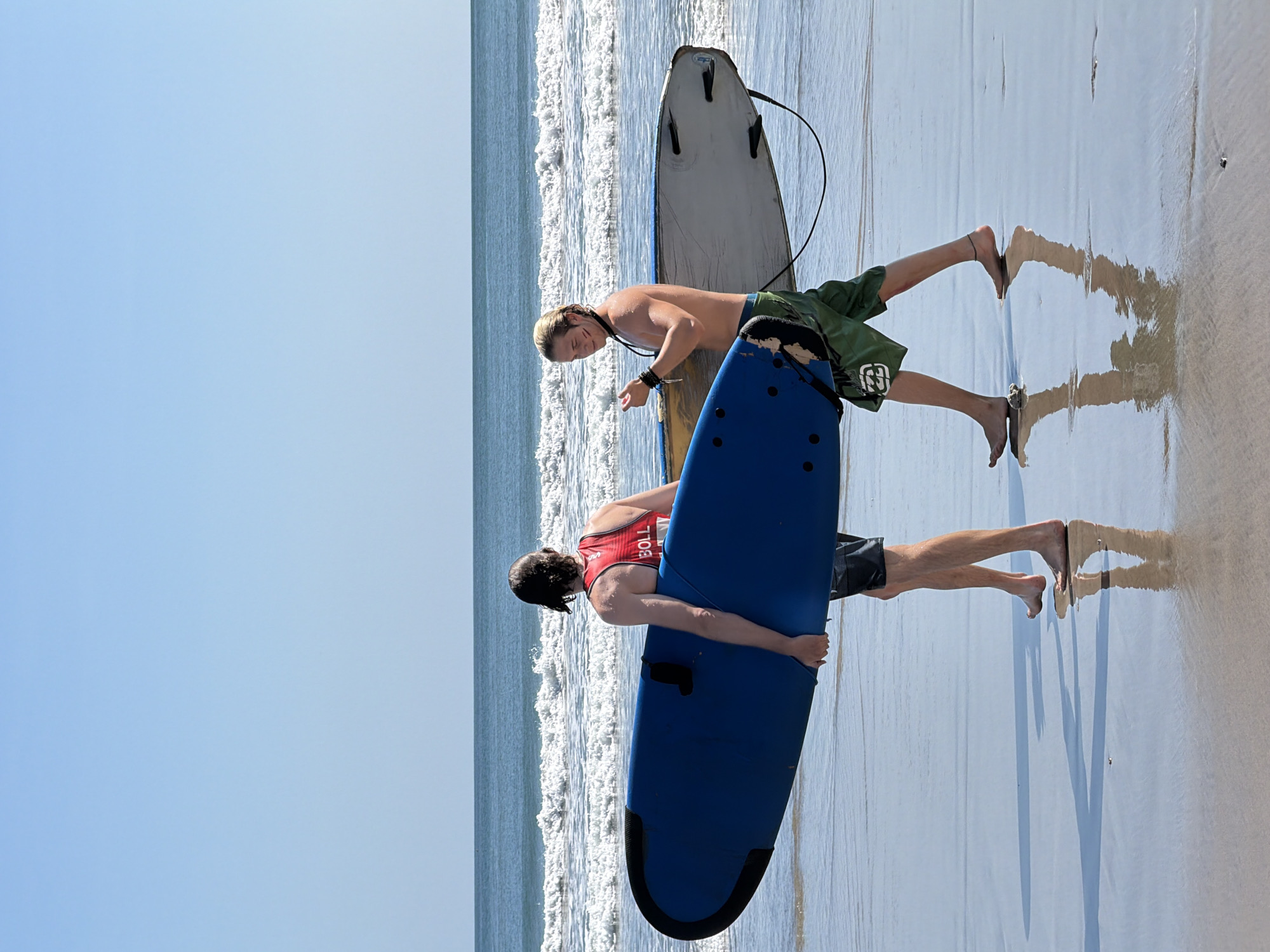 Two shirtless men are walking on a wet sandy beach, carrying surfboards. The man on the left is wearing a red tank top and dark shorts. He is holding a large blue surfboard. The man on the right is wearing green shorts and has his arm extended, holding a lighter colored surfboard. Both men are facing away from the camera and walking towards the right of the frame. The ocean with moderate waves is visible in the background, under a clear blue sky. The wet sand reflects the sky and the men, indicating the tide is out or has recently receded. The scene is consistent with the beaches of Kuta, Indonesia, known for surfing.