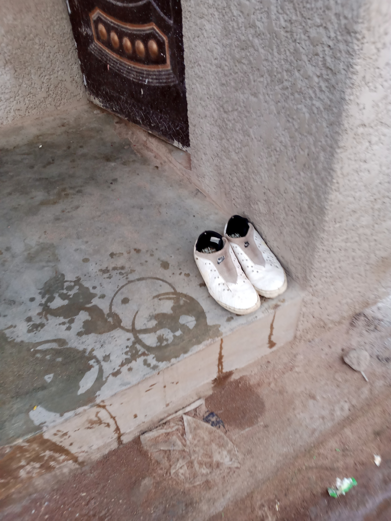 In Jauro Yinu, Nigeria, a pair of white slip-on shoes with black inner linings and tan Nike emblems rests on a concrete step. The shoes are positioned side-by-side, with the left shoe slightly forward. The concrete step is wet, with visible water stains and circular marks that suggest previous spills. To the left of the step, the ground is also wet and discolored. Behind the step, a textured concrete wall runs vertically. To the far left, a portion of a dark-colored door with ornate, metallic embellishments is visible. The surrounding ground is a mix of dirt and gravel, with some dry leaves and debris scattered about. The scene appears to be an outdoor entryway.