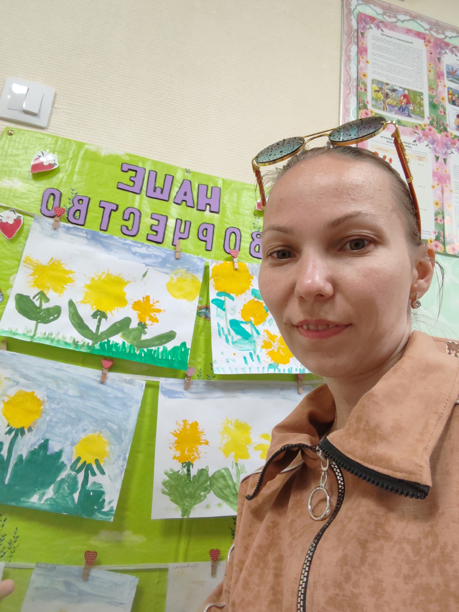 A woman stands in front of a bulletin board displaying children's artwork in Чухлэм, Russia. The board is bright green with a decorative border at the top and the backward text "НАШИ ТВОРЧЕСТВО" (Our Creativity) in purple. The children's drawings depict yellow flowers and green stems and are held in place with small clothespins with red hearts. The woman has her sunglasses placed on her head. She is wearing a collared, zippered jacket. Another poster with a floral border is partially visible on the upper right of the frame. A light switch is on the upper left.