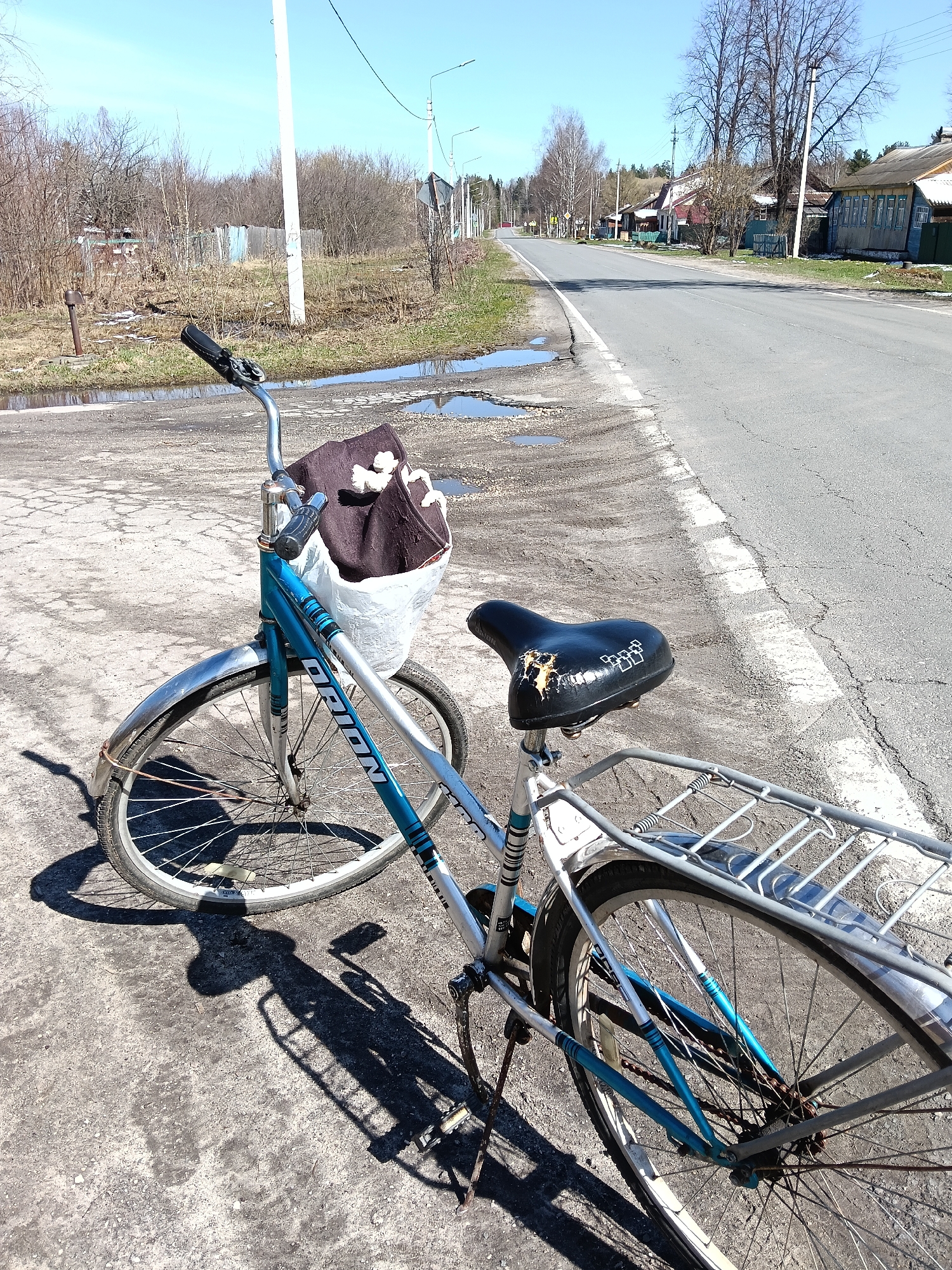 A blue and silver "ORION" bicycle is parked on a dirt and gravel shoulder next to an asphalt road, serving as the central subject of the image. A white basket attached to the handlebars holds a brown fabric item and some lighter objects. The black bicycle seat is visibly worn and torn, featuring a pattern of white squares. The bicycle also has a silver rear rack and fenders, with a somewhat rusty chain.
The scene depicts a quiet, semi-rural setting, consistent with a village in Красный Маяк, Russia. The asphalt road is cracked and dotted with potholes, some containing puddles, suggesting recent rain or snowmelt. The ground on the shoulder is also wet and uneven. In the background, numerous bare trees and bushes, along with patches of early green grass, indicate early spring. Traditional wooden houses of various colors line the road in the distance, interspersed with more trees, utility poles, and power lines under a clear, bright blue sky.
Sharp shadows cast by the bicycle suggest it is midday or early afternoon on a sunny day. No people are visible, creating a serene and ordinary atmosphere. The only discernible text is the brand name "ORION" on the bicycle's frame.
