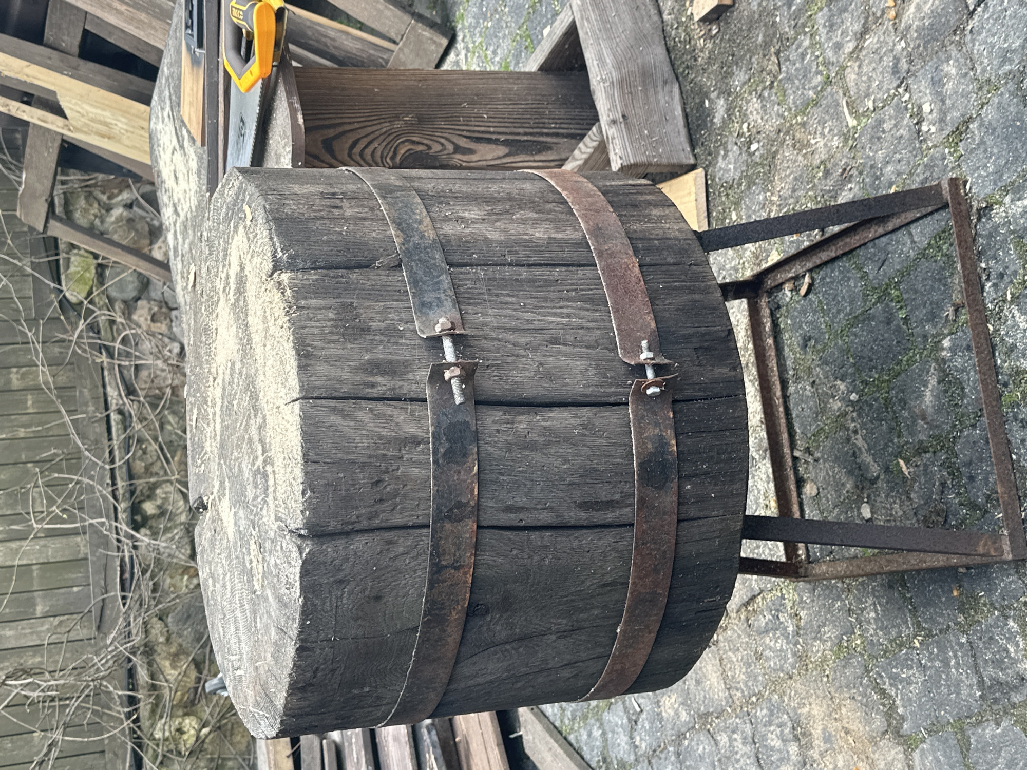 The image depicts a section of a large wooden log, secured with two metal bands that are fastened with bolts. The log rests on a rusted metal stand, which appears to be placed on a cobblestone surface. In the background, there is a partially visible wooden structure and some foliage, including small branches. A yellow-handled saw is visible in the upper left of the image. The scene is located in Новоглаголево, Russia.
