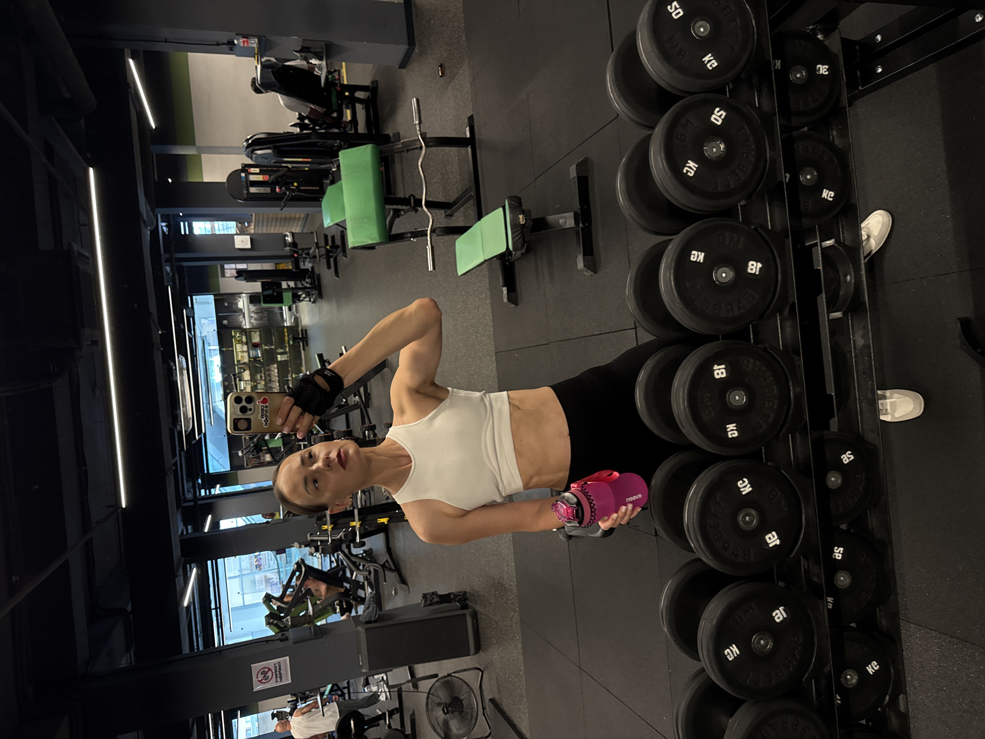A woman with brown hair pulled back is taking a mirror selfie in a gym. She is wearing a white crop top, black leggings, and black weightlifting gloves. She is holding a pink water bottle in her left hand and her right hand is raised to take the photo. In the background, there are various gym machines, including weight benches and dumbbell racks. The gym appears to be well-lit with fluorescent lights on the ceiling. The floor is made of black rubber matting. There are other people visible in the background, using gym equipment. The location is a gym, likely in a city like Sochi, Russia, as indicated by the language on some of the gym equipment.