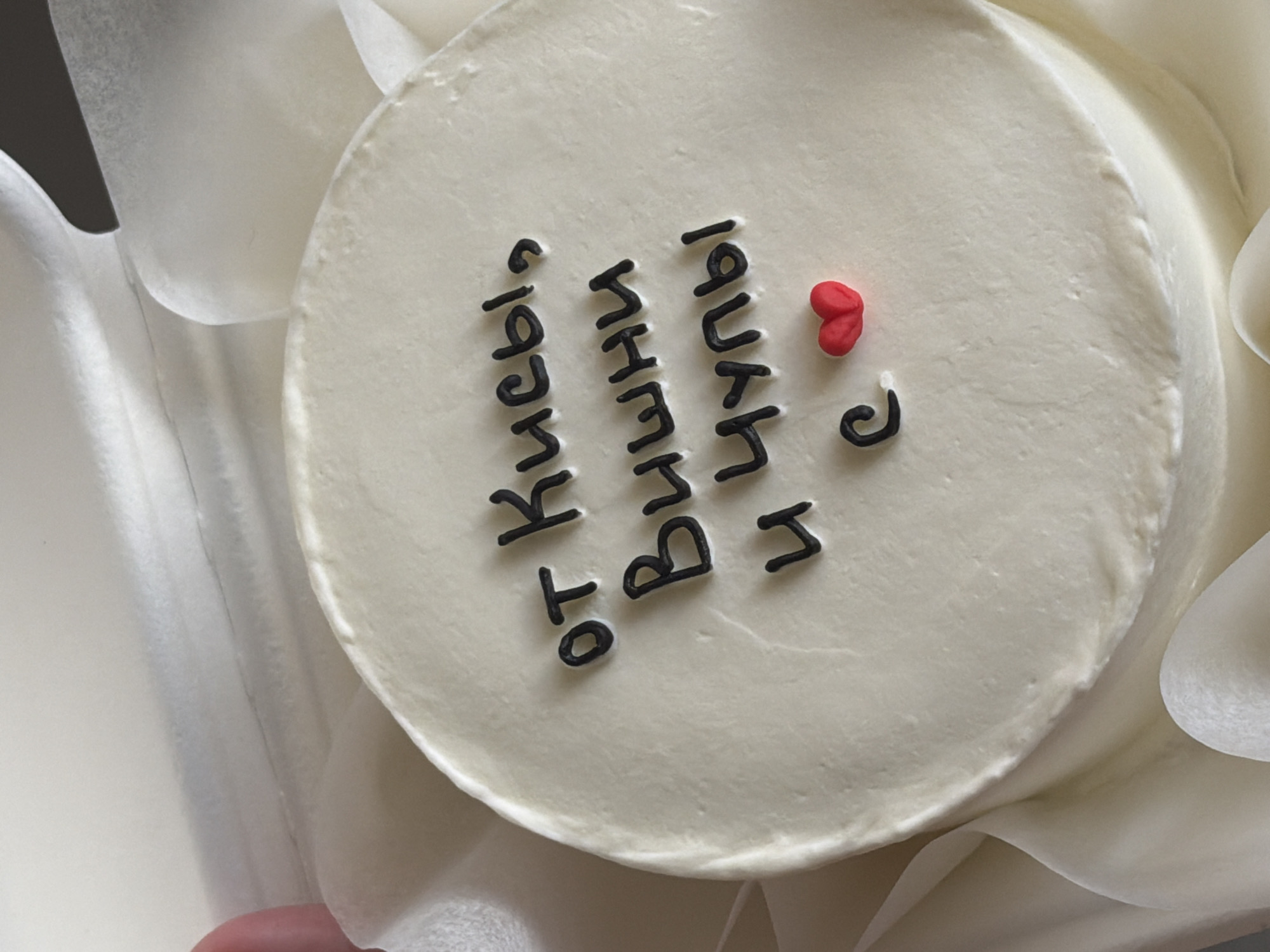 A close-up, overhead shot shows a white frosted cake with black frosting text and a small red heart decoration. The text appears to be in Cyrillic script and reads "от Кисьы, Вишни и Чупы с" which translates to "from Kisy, Cherries and Chupy with". The cake is sitting on a surface covered with white fabric, possibly tissue paper or a tablecloth, which is gathered around the edges of the cake, creating a soft, diffused background. The lighting is soft and even, highlighting the texture of the frosting. No specific location in Tel-Aviv, Israel is discernible from the image.