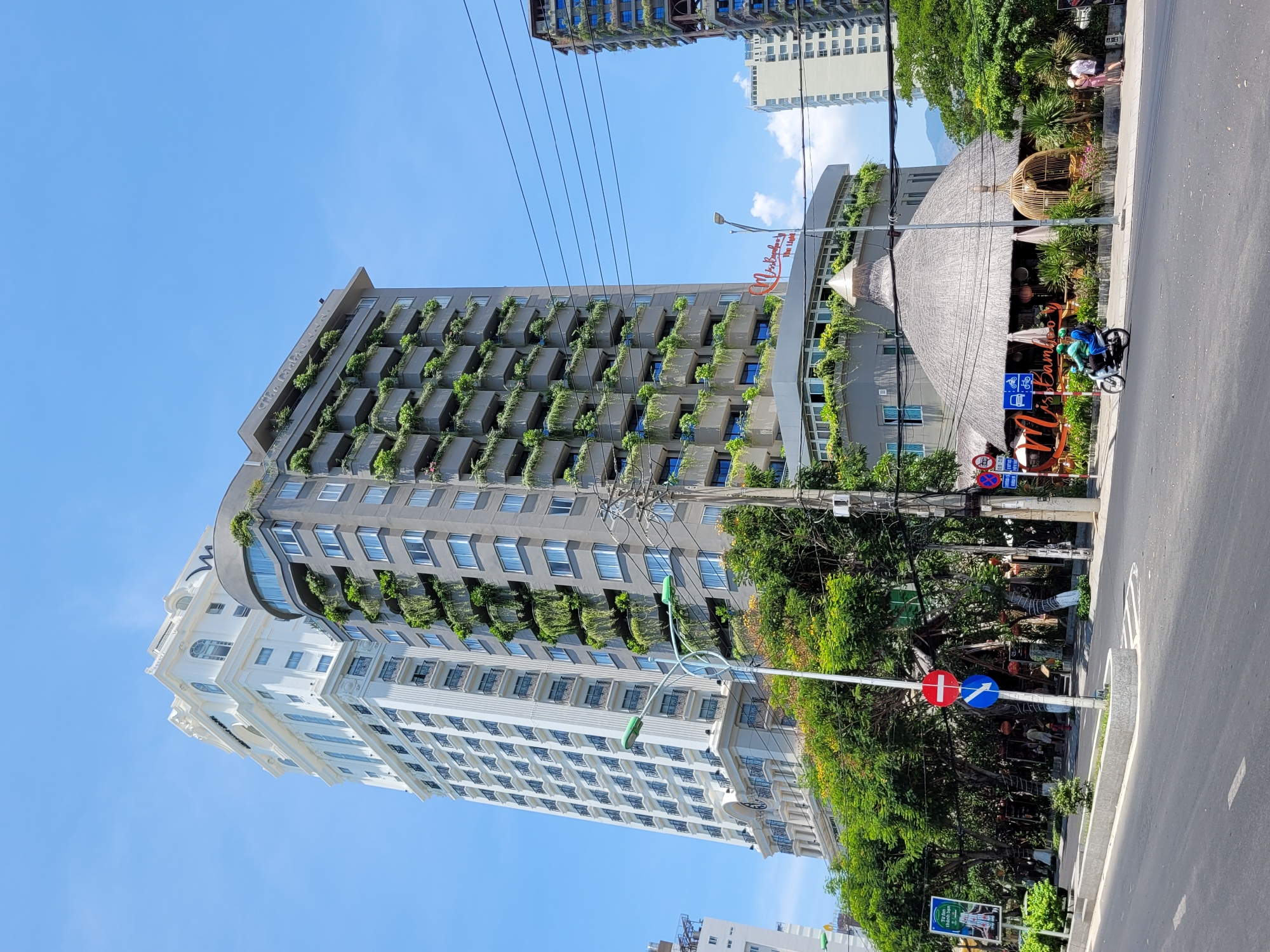 The photograph captures a multi-story building with gray and white architectural features. Balconies with greenery adorn the gray sections of the building. A restaurant or establishment with a thatched roof and signage is adjacent to the building. A street is visible with a person riding a motorbike. Street signs are present. The sky is clear blue.
