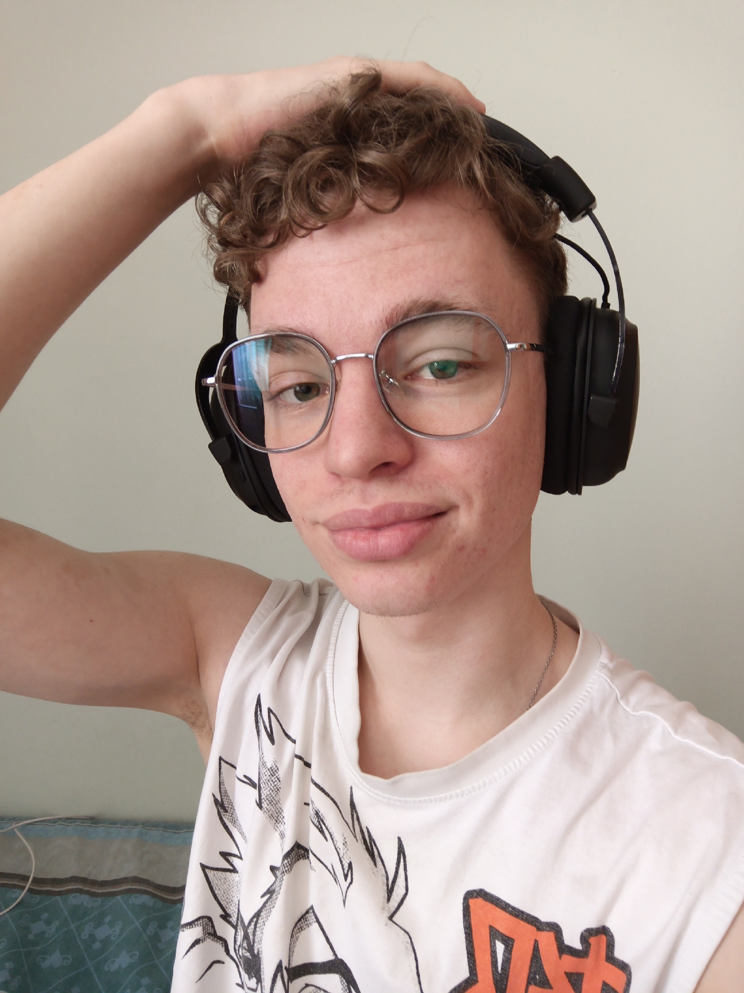 A young man is taking a selfie in what appears to be an indoor setting. He is wearing black headphones over his head and silver-rimmed glasses. His hair is short and curly. He is wearing a white tank top with a graphic design. His left hand is on top of his head. He has fair skin and a slight smile. The background is a plain, light-colored wall. A cord is visible along the bottom left of the frame. There is a glimpse of a bed with a patterned blue and gray blanket. There is no indication of the location being in Odintsovo, Russia.