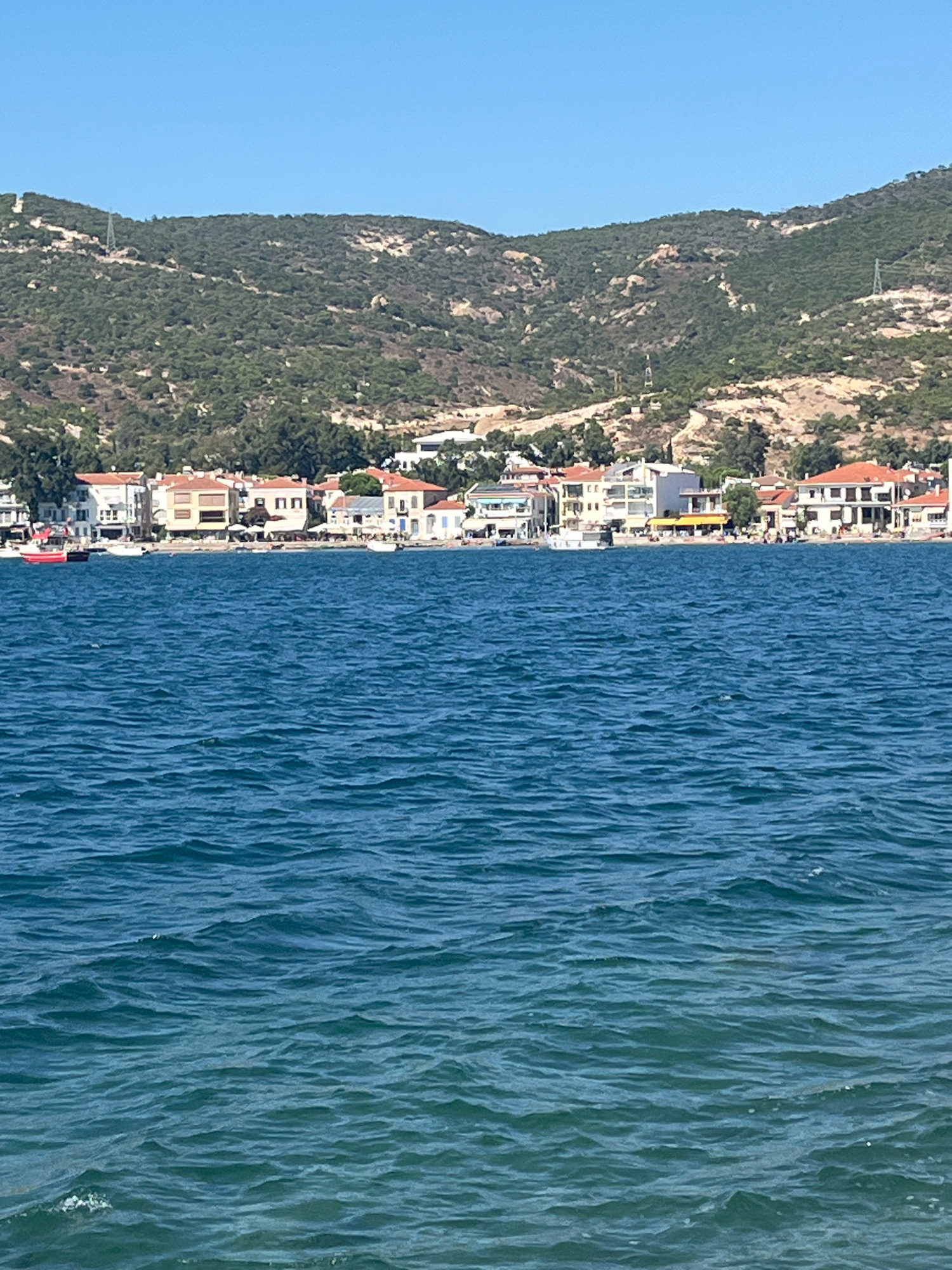 The image shows a body of water with a town and hills in the background, likely Foça, Turkey. The foreground is dominated by rippling blue water. In the midground, a line of buildings with red-tiled roofs and white facades runs along the shore. Several boats are visible near the shore, including a prominent red boat. Behind the town, green, scrub-covered hills rise up, with some evidence of terracing or development. The sky is clear and blue. There are no people clearly visible in the image. The overall scene depicts a coastal town on a sunny day.