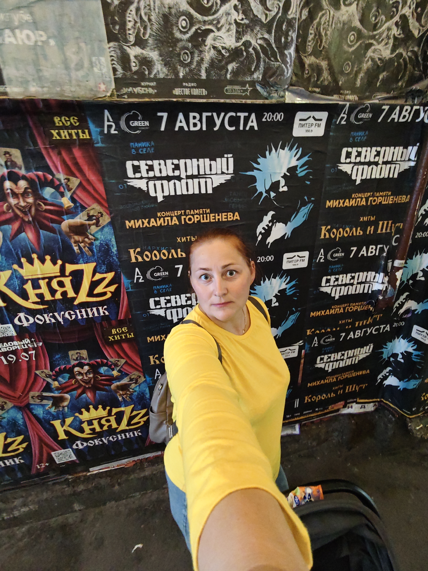 The image shows a woman in a bright yellow shirt and blue jeans taking a selfie in front of a wall covered with posters in Saint Petersburg, Russia. The posters advertise concerts, including "КняZz Фокусник" (KnyazZ Juggler) with a date of 19.07, and "Северный Флот" (Northern Fleet) in memory of Mikhail Gorshenev. There are also smaller posters with text in Russian and logos for Green Concert and Piter FM. The environment appears to be an indoor space, possibly a venue or a backstage area, with a slightly grimy floor. The woman is positioned in the center of the frame, looking directly at the camera with a slightly surprised expression. A stroller is partially visible to her right.