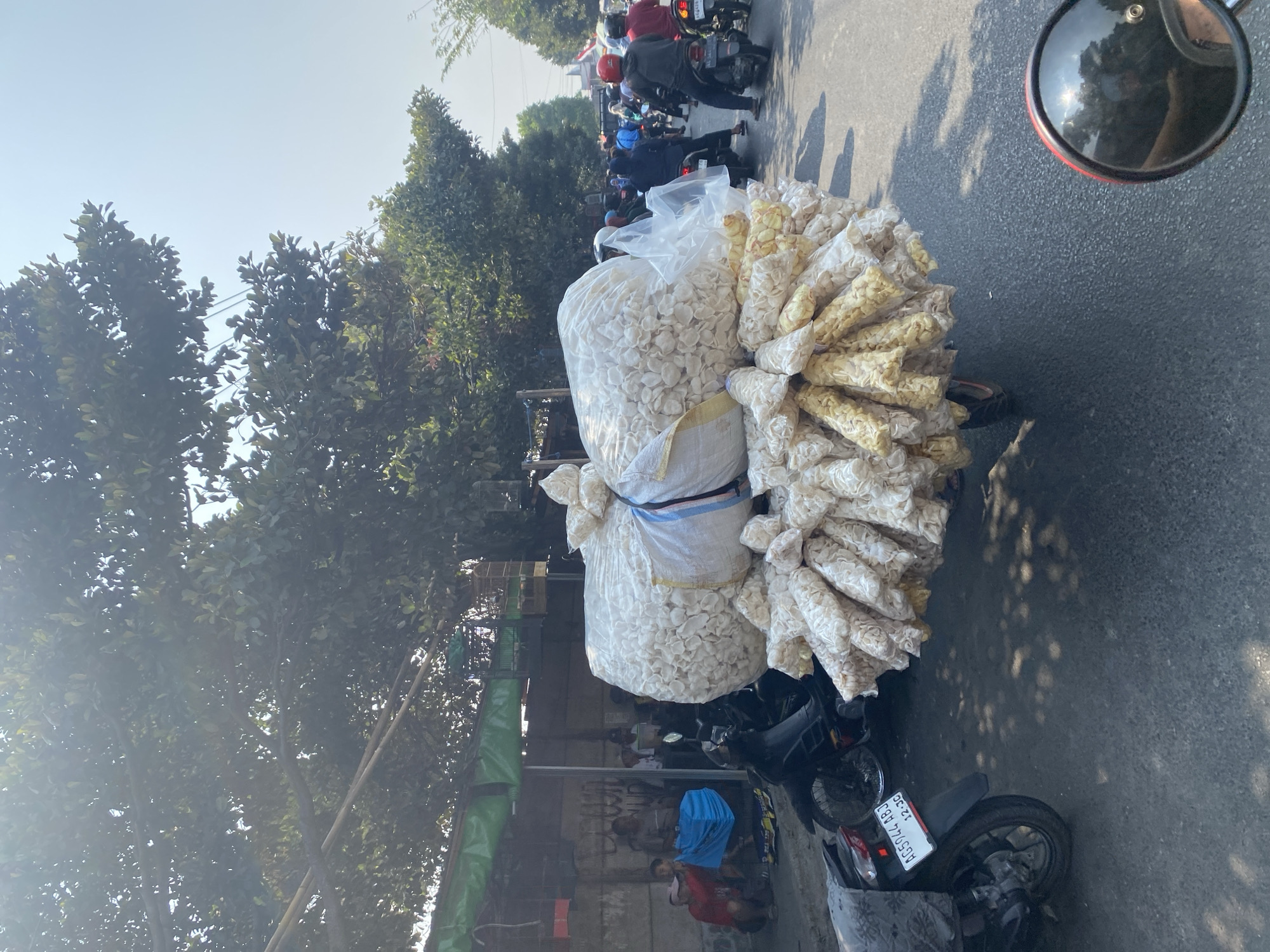 In Kediri City, Indonesia, on a sunlit day, a large cart laden with packaged snacks is being transported along a street. The cart is piled high with numerous transparent plastic bags, each containing white, puffed snack items, some appearing cylindrical and others flatter. A large white sack, also filled with similar snacks, sits on top of the cart. In the background, several people are visible near a row of shops, some sitting and others standing. Motorcycles are also present, with one parked in the foreground and others in motion on the street, indicating a busy urban environment. The scene suggests a vendor delivering goods or a market setting.