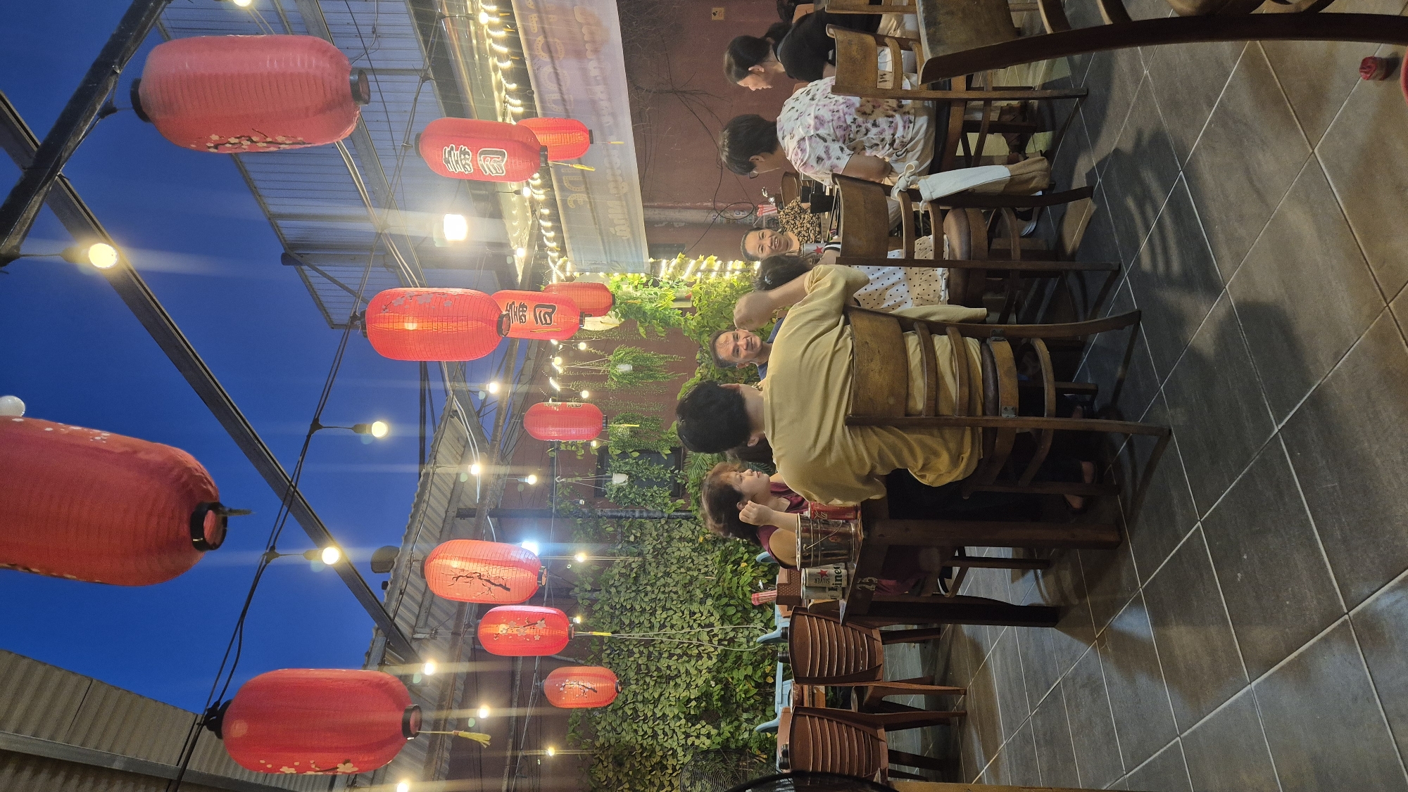 A group of people are gathered at tables in an outdoor restaurant or dining area, likely in Đà Nẵng, Vietnam, under a twilight sky illuminated by numerous red lanterns. The restaurant features a roof structure with exposed beams and metal supports from which strings of lights and the lanterns hang. Lush green foliage cascades from above and covers the background walls. People are seated around wooden tables, some are in conversation, while others appear to be eating. The flooring is tiled in a dark grey grid pattern. The overall atmosphere suggests a casual evening meal.