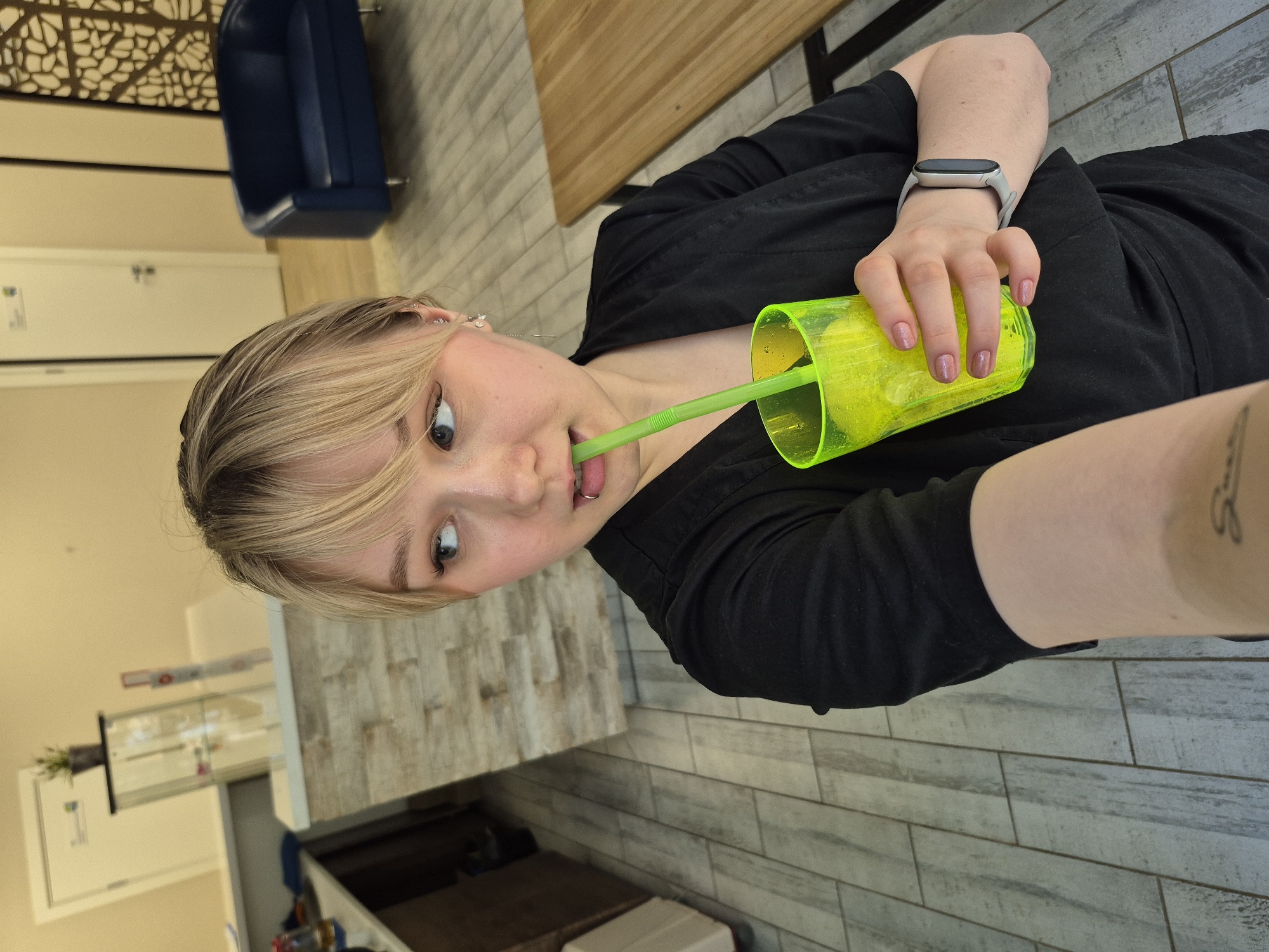 An indoor, eye-level, slightly rotated image captures a young person with blonde hair and bangs looking directly at the camera. They are wearing a black short-sleeved top and have a playful, wide-eyed expression with their mouth slightly open, a neon green straw in it, and their tongue visible. A small silver ring piercing is present on their lower lip. Their nails are painted a light, shimmery pink, and a light grey fitness tracker or smartwatch is worn on their left wrist. A tattoo is partially visible on their right forearm.
The person is holding a translucent neon green cup filled with ice and a beverage, with a matching green straw. The background features light-colored walls and a floor with rectangular tiles resembling pale wood planks. Elements visible in the background include a dark blue upholstered chair, a light wooden table or counter, and a decorative wall panel with an abstract, organic pattern. Further back, a white door and a clear display case or shelf with small items, possibly plants, are discernible. The setting appears to be a modern indoor space, potentially a cafe, lounge, or waiting area in Chaikovsky, Russia. No discernible text is visible.